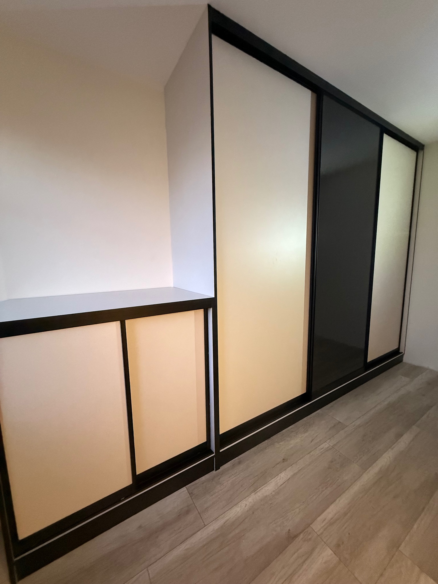 Nowoczesna szafa wnękowa z przesuwnymi drzwiami w odcieniach beżu i czerni, z dodatkową półką po lewej stronie, na tle jasnej podłogi.