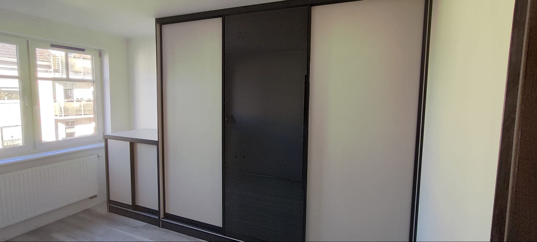 Nowoczesna szafa wnękowa z przesuwnymi drzwiami w odcieniach bieli i grafitu, z dodatkową półką boczną, w jasnym wnętrzu z oknem.