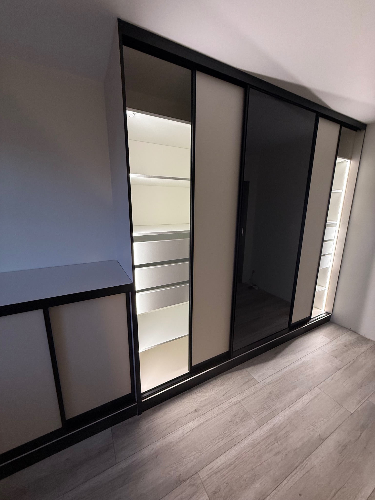 Szafa wnękowa z przesuwnymi drzwiami, beżowo-czarna, z oświetleniem LED wewnątrz, widok z boku, na podłodze panele imitujące drewno, nowoczesny design.