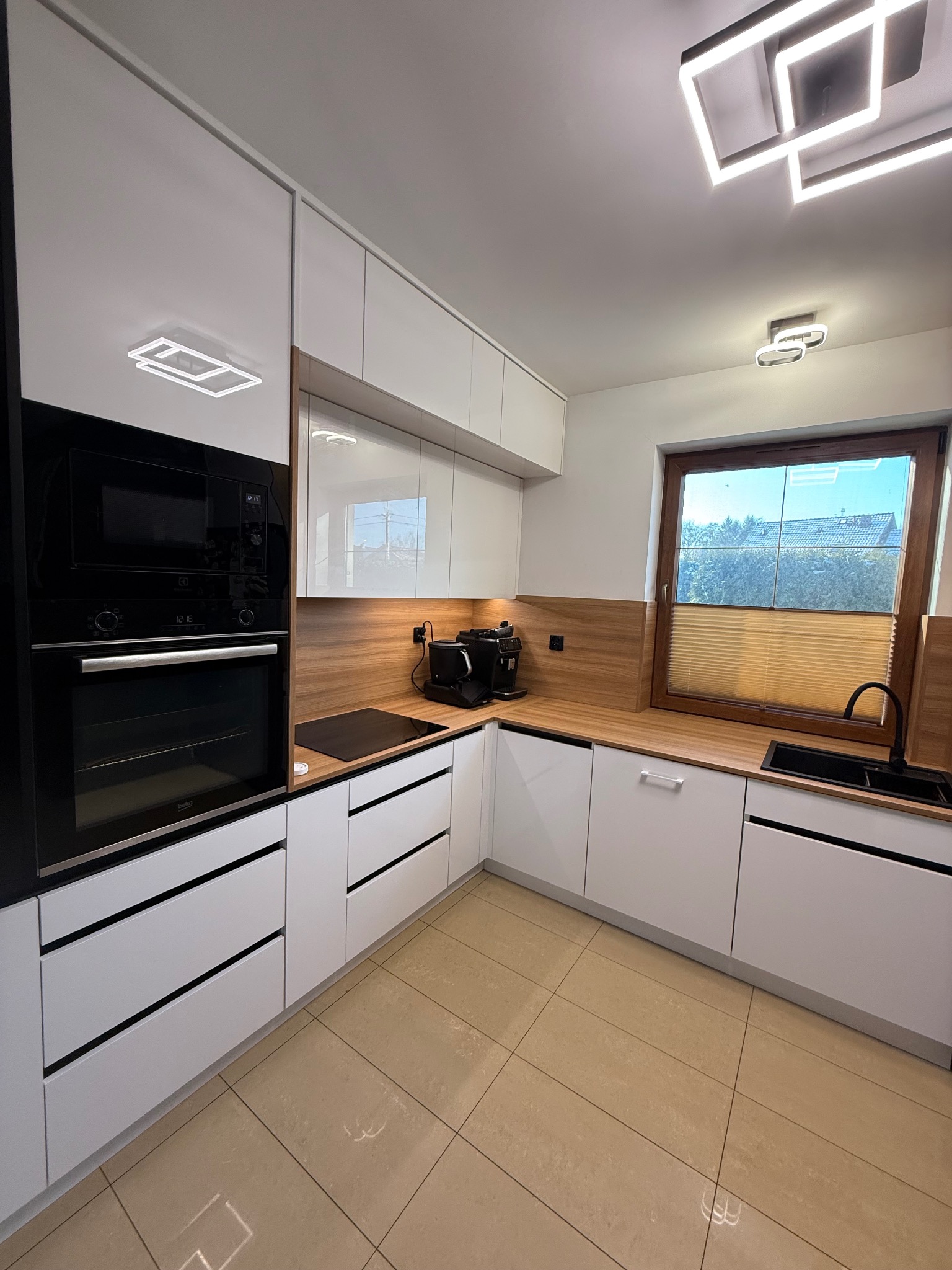 Nowoczesna kuchnia z białymi, lakierowanymi frontami, drewnianym blatem i czarnymi akcentami: piekarnik, płyta indukcyjna i zlew. Oświetlenie LED sufitowe.