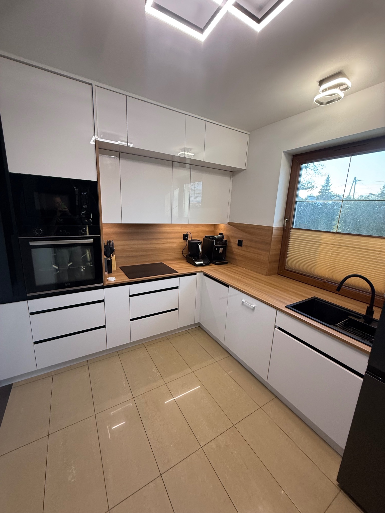 Nowoczesna kuchnia z białymi frontami, drewnianym blatem i czarnymi akcentami, w tym zlew i bateria. Widok na okno z żaluzjami.