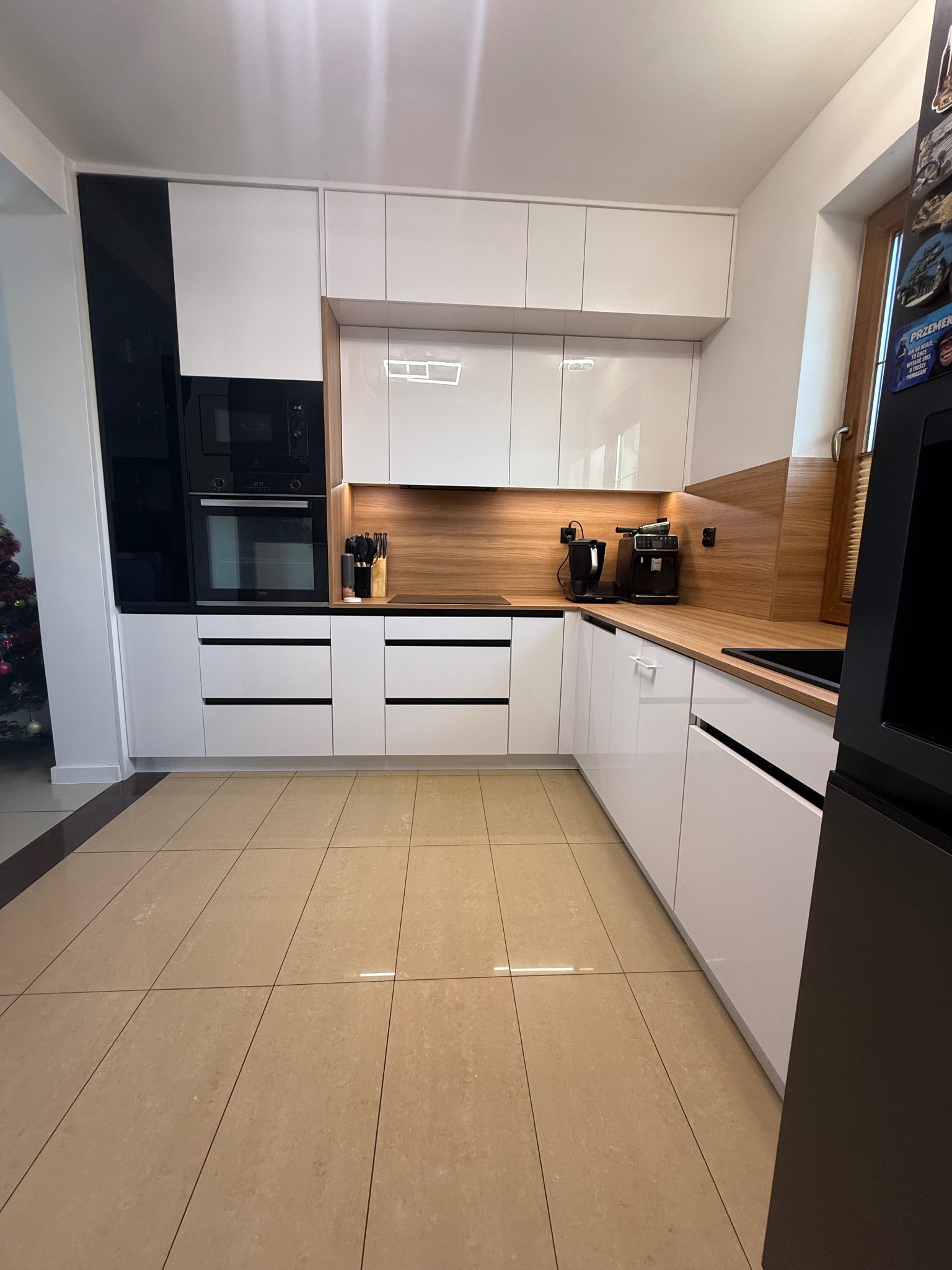 Nowoczesna, biała kuchnia z drewnianym blatem i czarnymi akcentami. Widoczny sprzęt AGD i oświetlenie podszafkowe. Na podłodze beżowe płytki. W tle choinka.