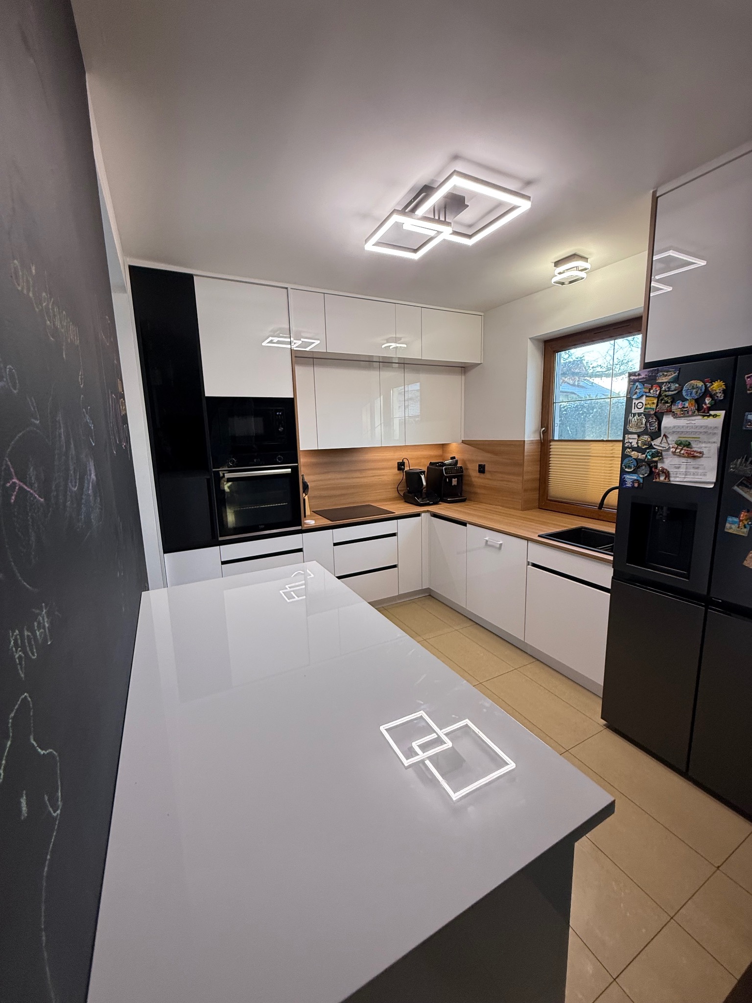 Nowoczesna kuchnia z białymi szafkami, drewnianym blatem i czarnymi akcentami. Na pierwszym planie wyspa z podświetleniem LED. Widoczna tablica kredowa.