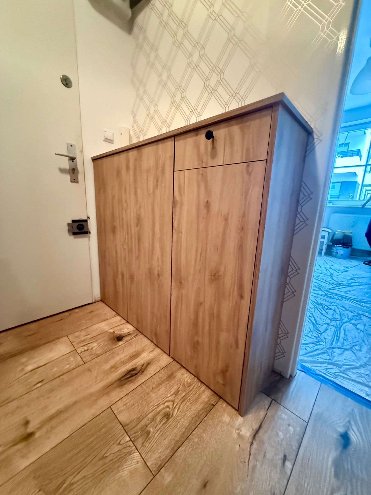 Drewniana szafka na buty z szufladą, dopasowana do przedpokoju z drewnianą podłogą i wzorzystą tapetą. Funkcjonalne rozwiązanie do przechowywania obuwia.