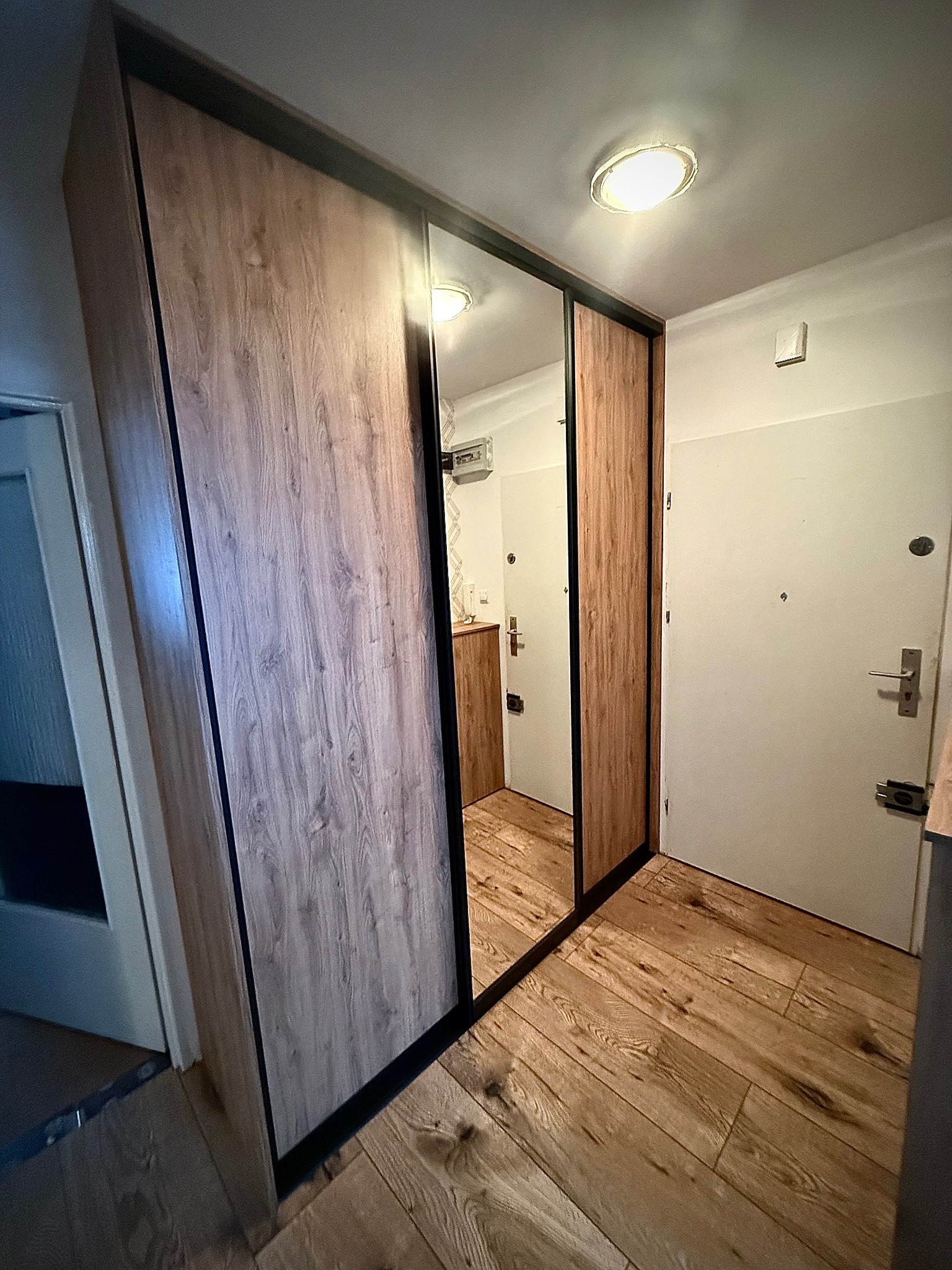 Wnękowa szafa z przesuwnymi drzwiami z lustrem i drewnopodobnym wykończeniem, wkomponowana w korytarz z drewnianą podłogą i białymi drzwiami.