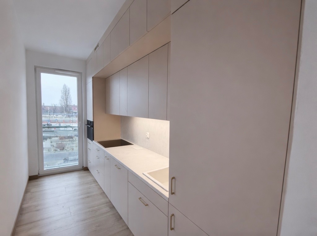 Jasna kuchnia na wymiar z zabudową po sufit, minimalistyczne fronty, złote uchwyty, oświetlenie LED pod szafkami wiszącymi, widok na zewnątrz przez okno.