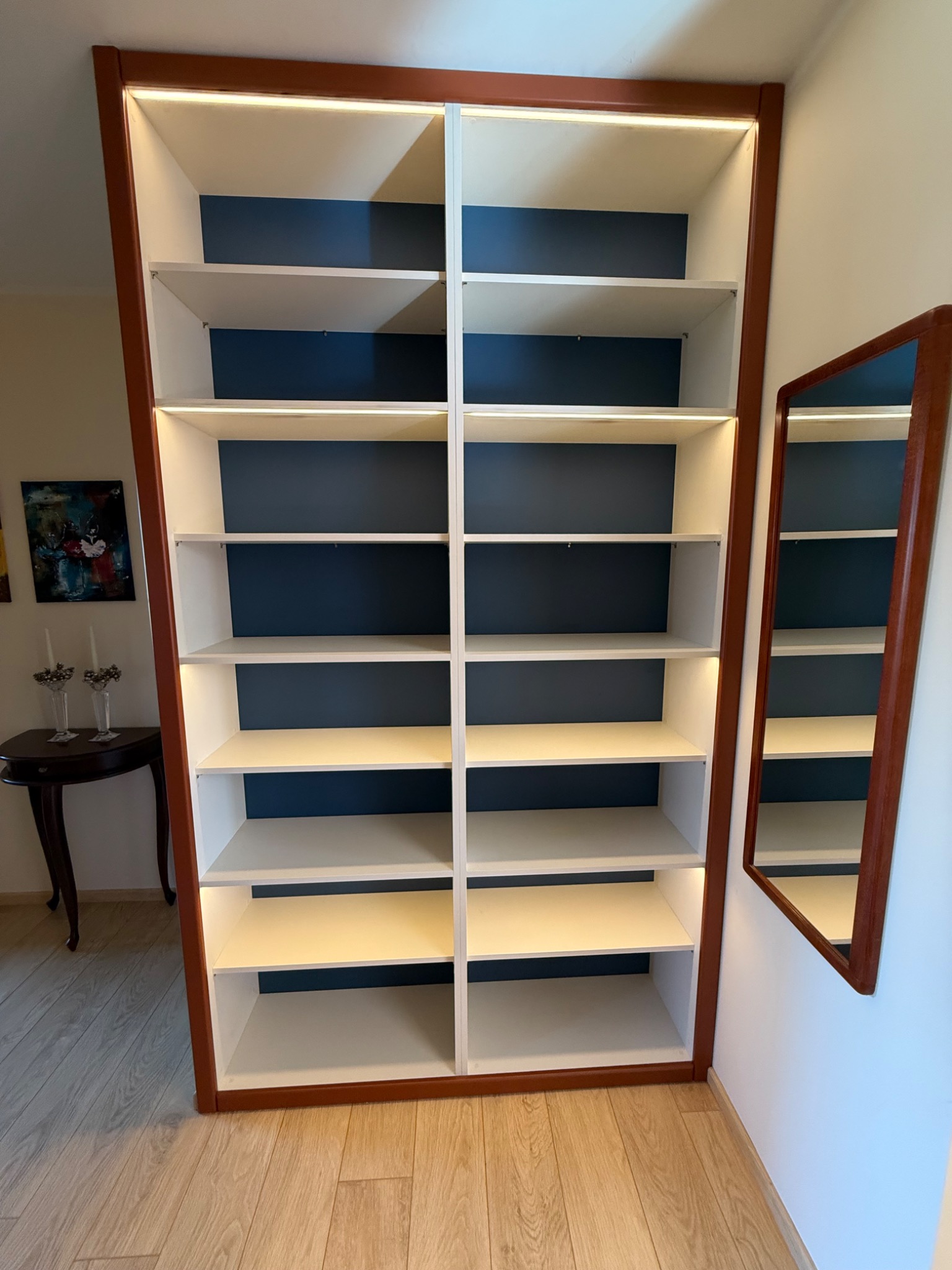 Wysoka, wnękowa szafa z podświetleniem LED, białe półki na tle granatowej ściany, obramowana brązowym drewnem. Obok lustro w pasującej ramie.