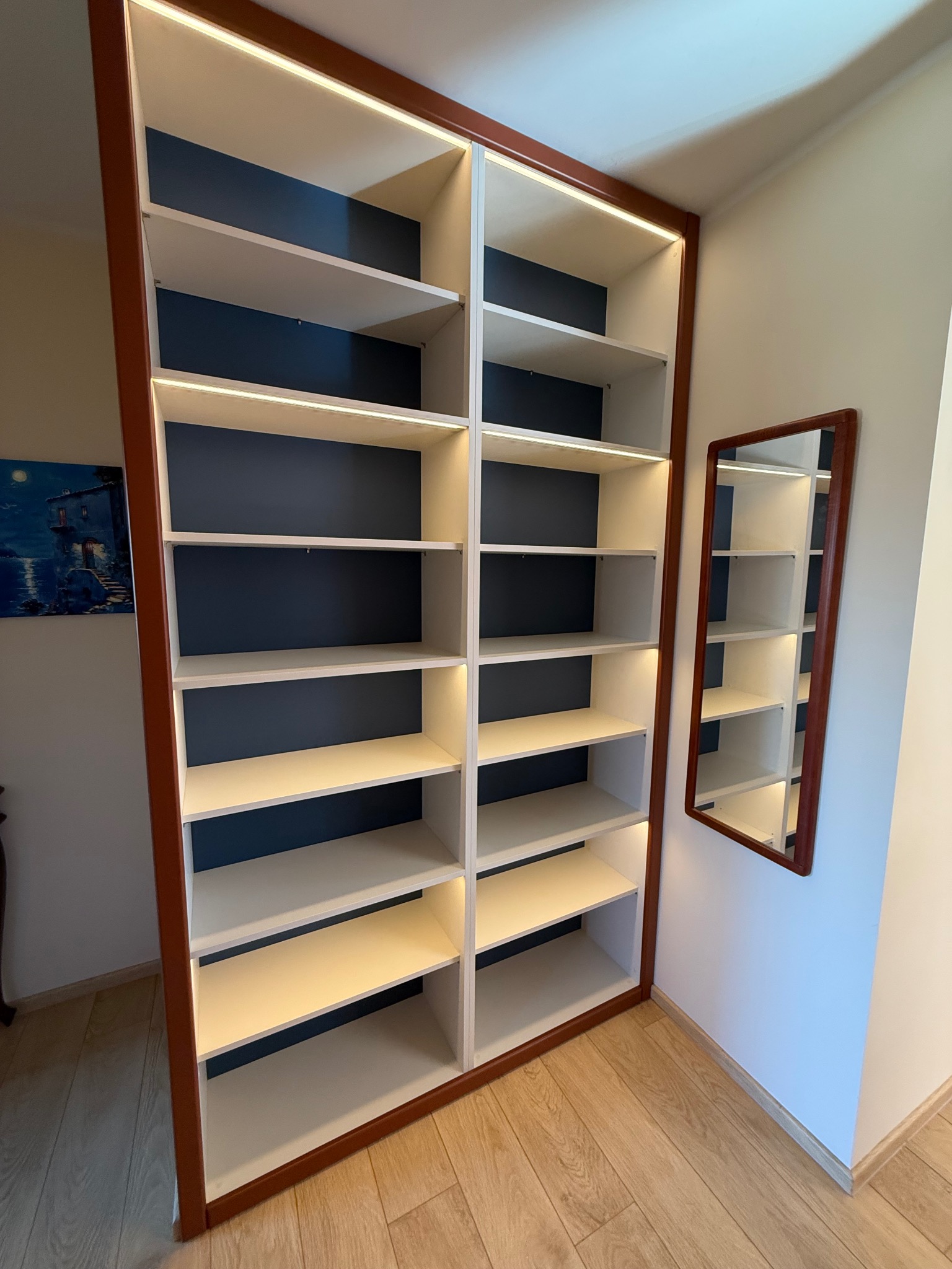 Wysoka, wnękowa szafa z podświetlanymi półkami i ciemnoniebieskim tłem, dopasowana do ściany. Obok lustro z ramą w tym samym kolorze co obramowanie szafy.