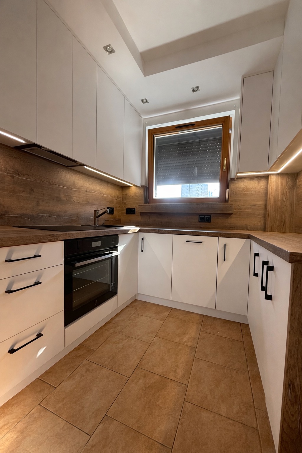 Nowoczesna kuchnia z białymi szafkami i drewnianymi blatami, oświetleniem LED podszafkowym i brązowymi płytkami podłogowymi. Minimalistyczne czarne uchwyty.