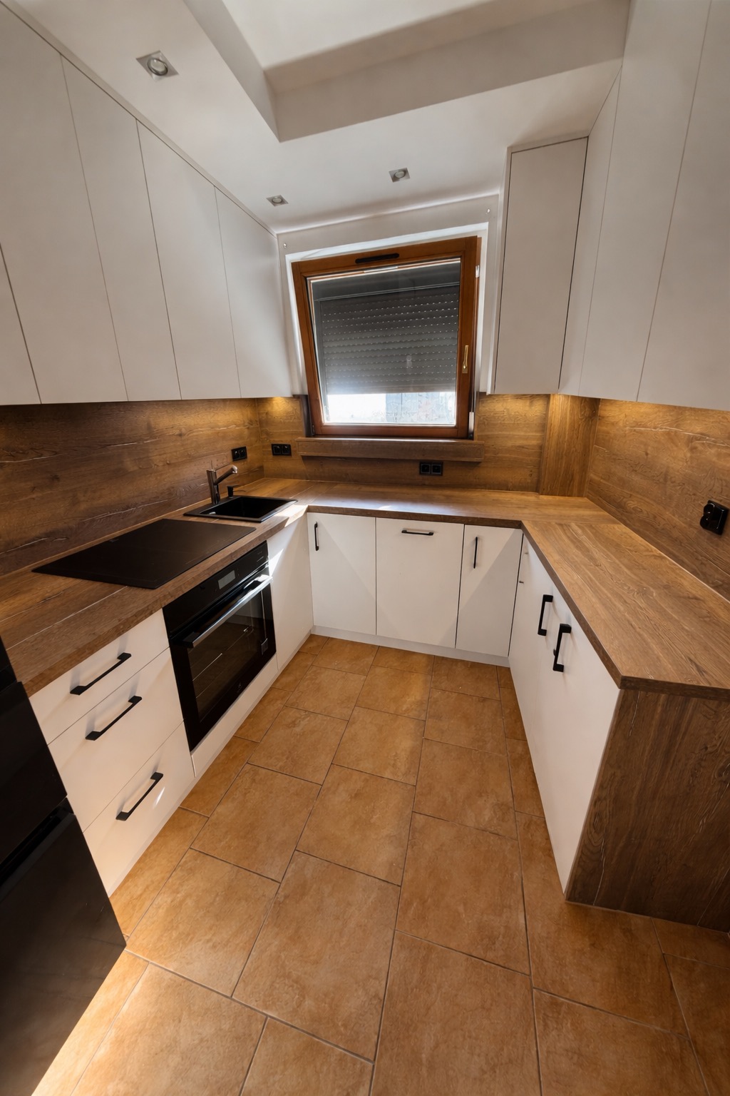 Nowoczesna kuchnia w kształcie litery U z białymi szafkami i drewnianymi blatami. Uwagę przykuwa okno z roletą i oświetlenie LED pod szafkami.