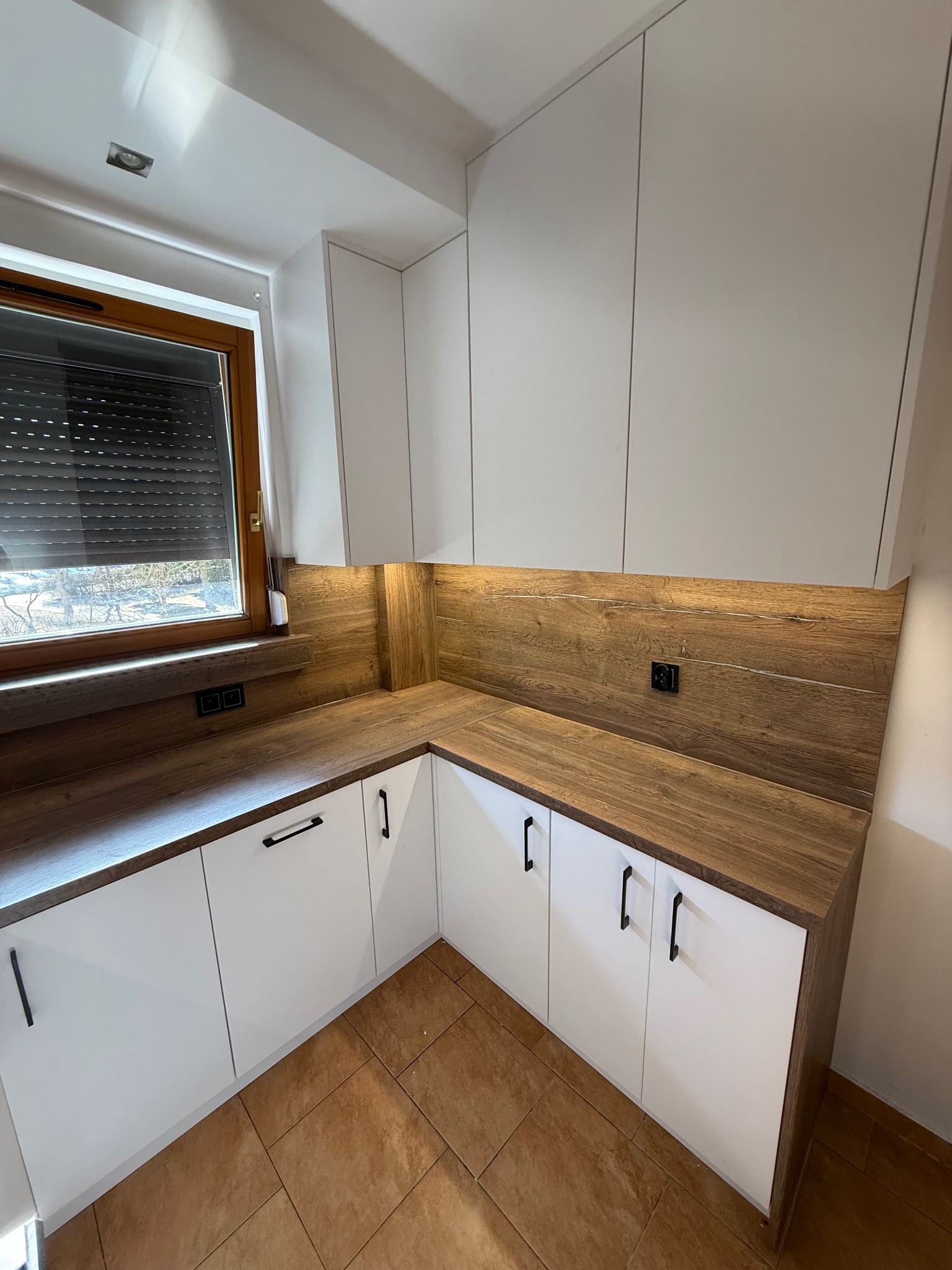 Białe szafki kuchenne z drewnianym blatem i oświetleniem LED pod szafkami wiszącymi, widok na okno z roletami i brązową podłogę.