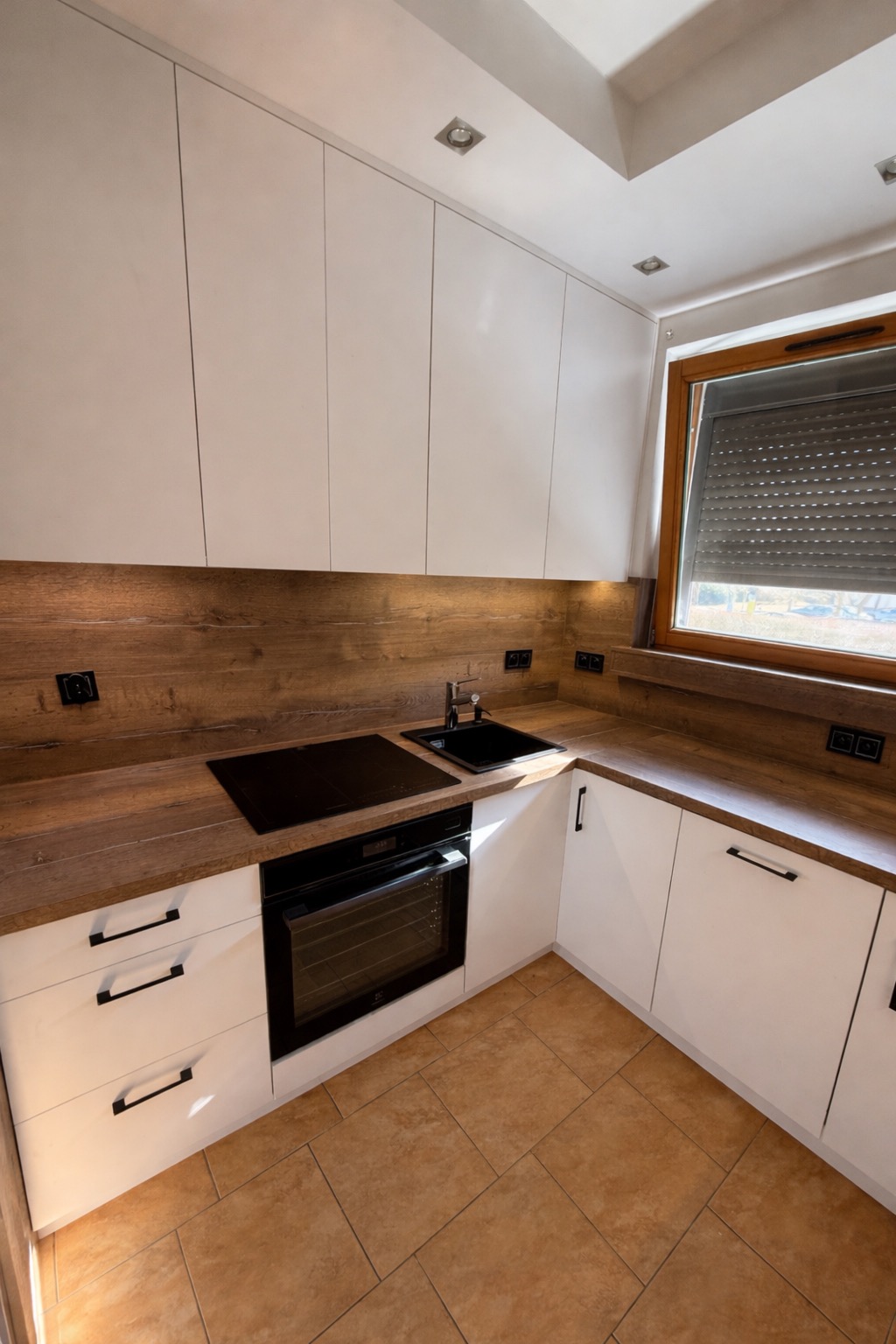 Nowoczesna kuchnia narożna z białymi szafkami i drewnianym blatem. Czarny zlew i płyta indukcyjna. Minimalistyczne uchwyty i ciepłe oświetlenie.