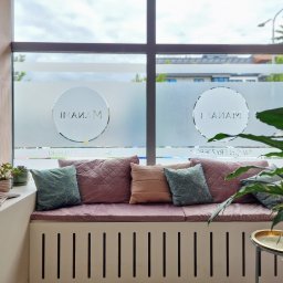 MANAMI Salon Fryzjerski Anna Dębska - Wnętrze z dużym oknem, na którym widnieje logo salonu, pod oknem znajduje się siedzisko z poduszkami, a po bokach rośliny doniczkowe i stolik.