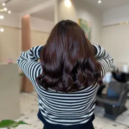 Tył głowy kobiety z brązowymi, błyszczącymi włosami ułożonymi w delikatne fale, w salonie z jasnym tłem i czarnym fotelem fryzjerskim.