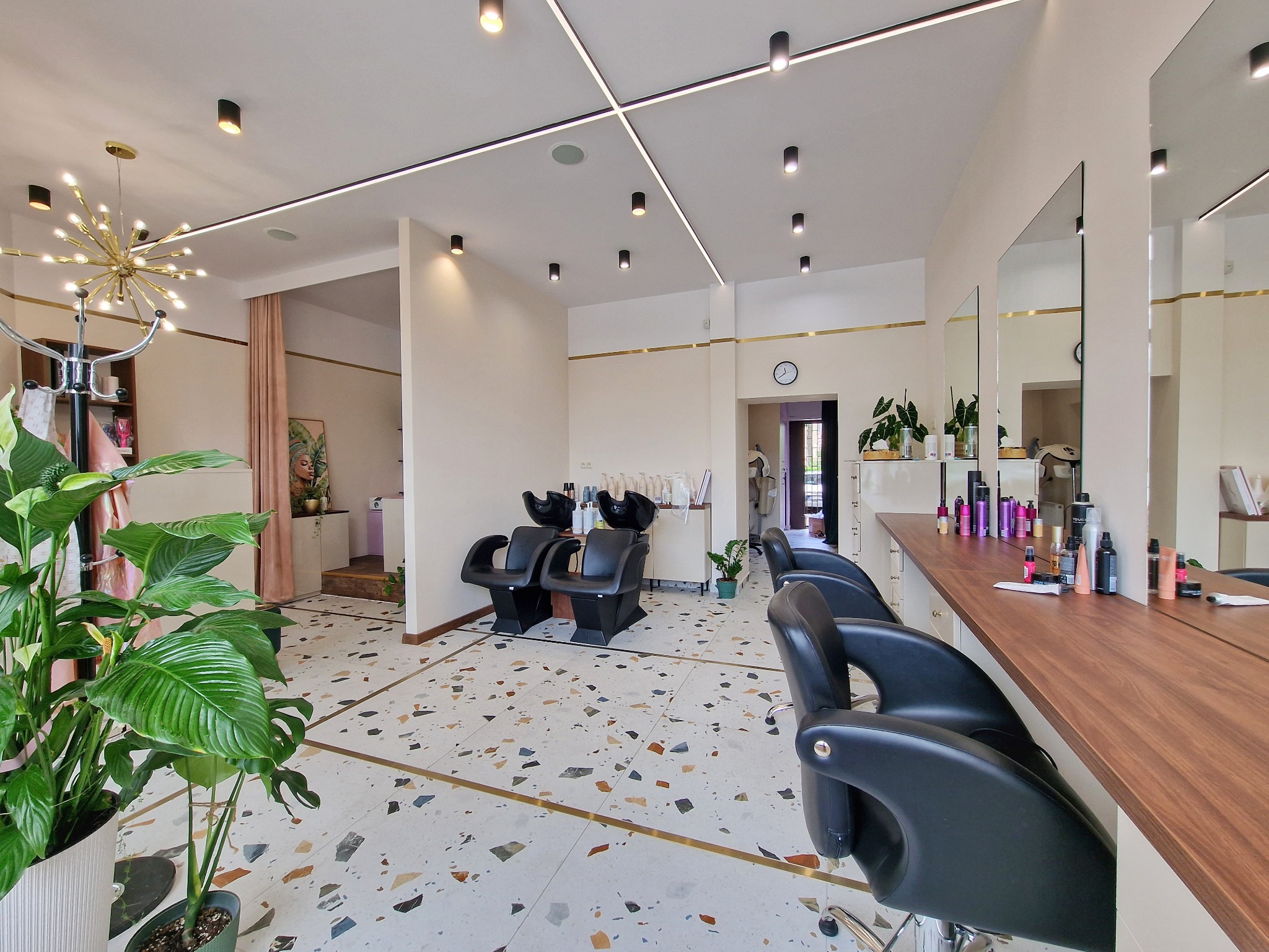 Jasne, przestronne wnętrze z terazzo, czarne fotele fryzjerskie, stanowiska z lustrami i kosmetykami, nowoczesne oświetlenie sufitowe oraz dekoracyjna lampa w stylu retro.