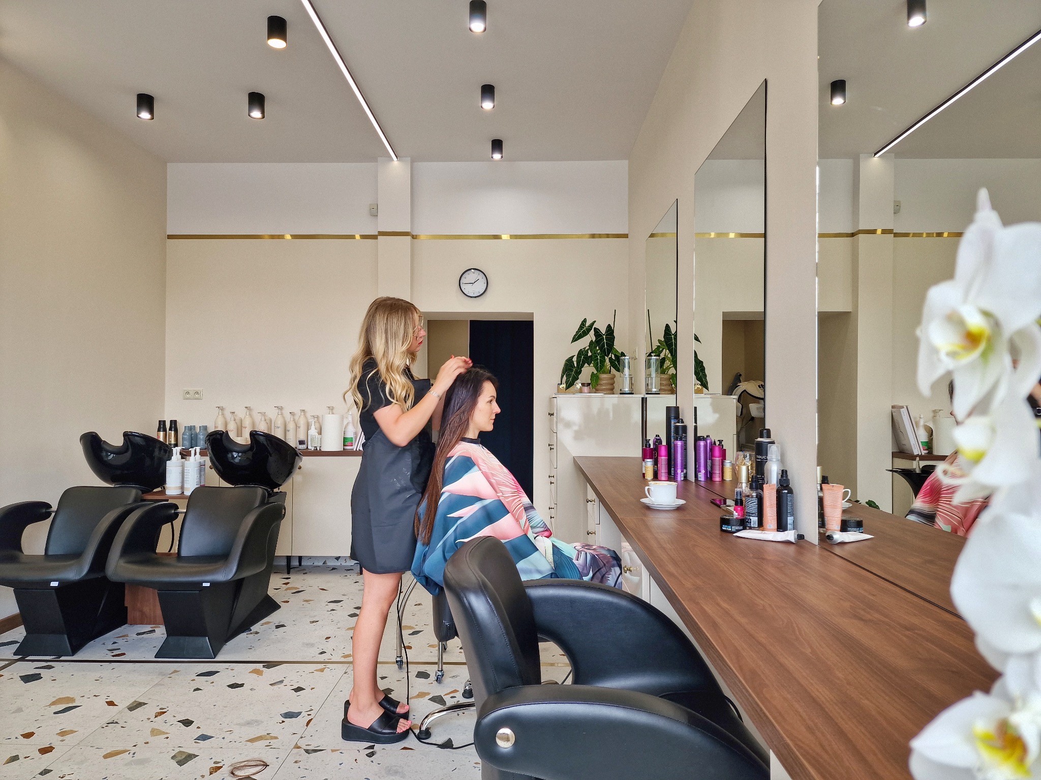 Kobieta w czarnym fartuchu, stojąca za siedzącą klientką z peleryną w salonie, poprawia jej włosy na tle minimalistycznego wnętrza z dużym lustrem i liniowym oświetleniem.