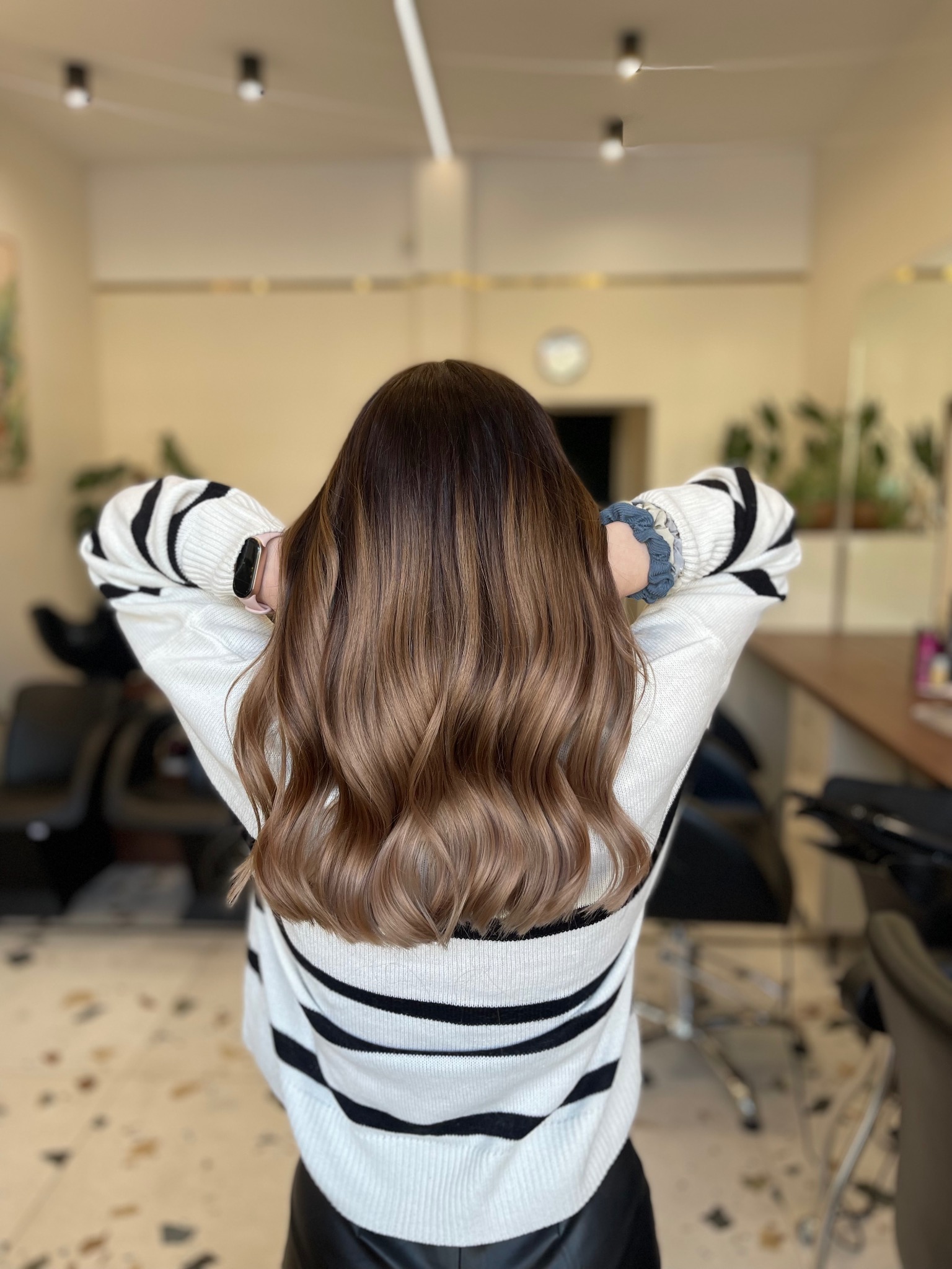 Tył głowy kobiety z włosami ombre w odcieniach brązu, delikatnie pofalowanymi, w salonie fryzjerskim, ubrana w sweter w czarno-białe paski, z gumką do włosów na nadgarstku.