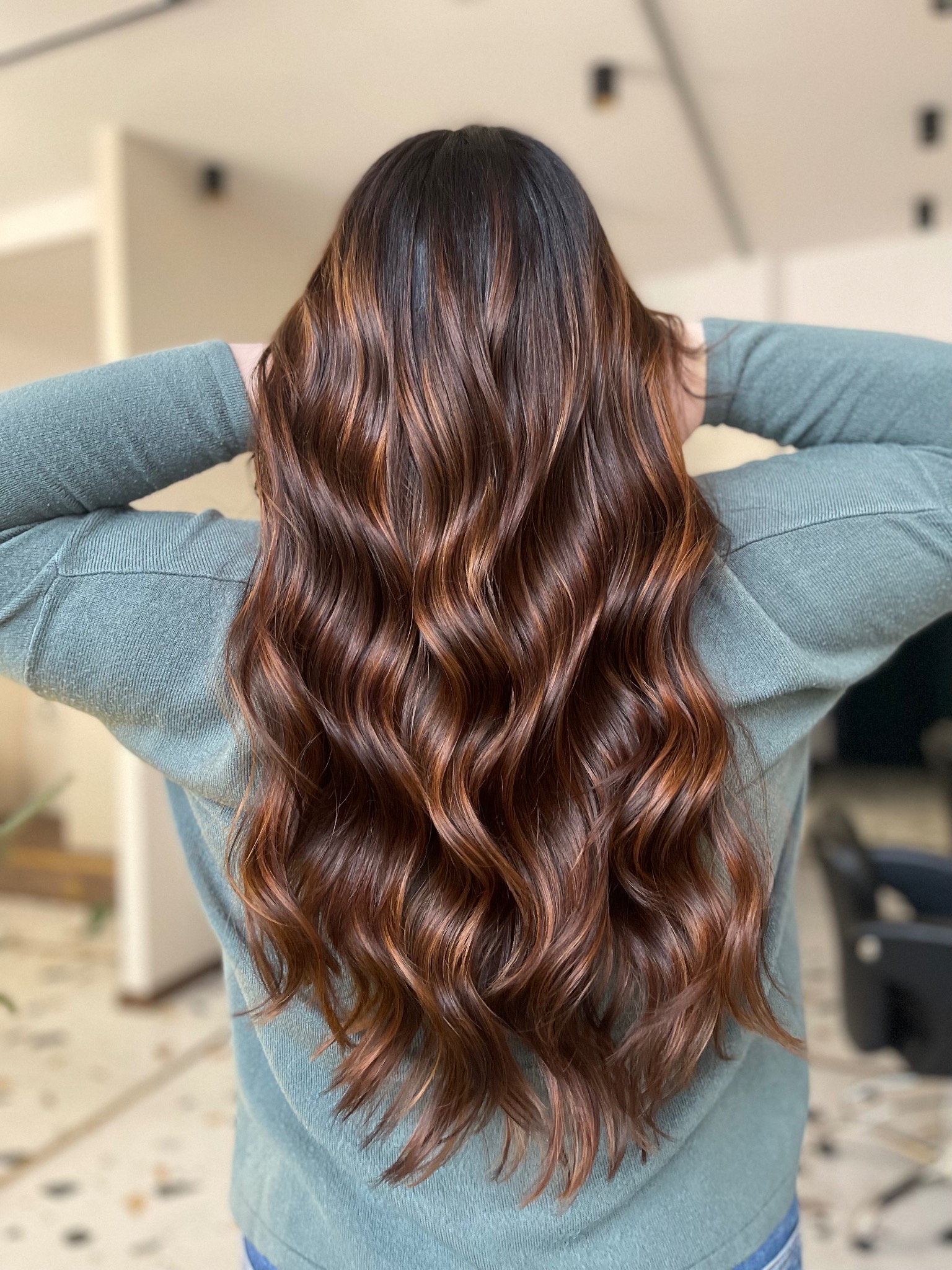 Długie, falowane, brązowe włosy z miedzianymi refleksami, widok z tyłu, kobieta w zielonym swetrze trzyma ręce na karku.