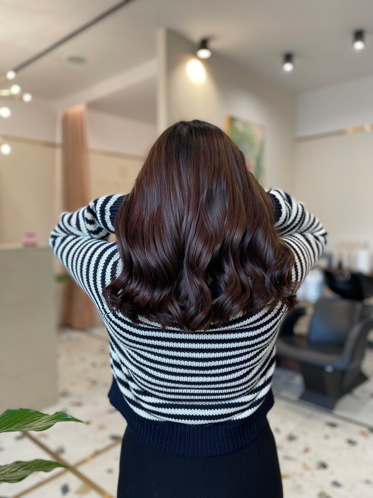 Tył głowy kobiety z brązowymi, błyszczącymi włosami ułożonymi w delikatne fale, w salonie z jasnym tłem i czarnym fotelem fryzjerskim.