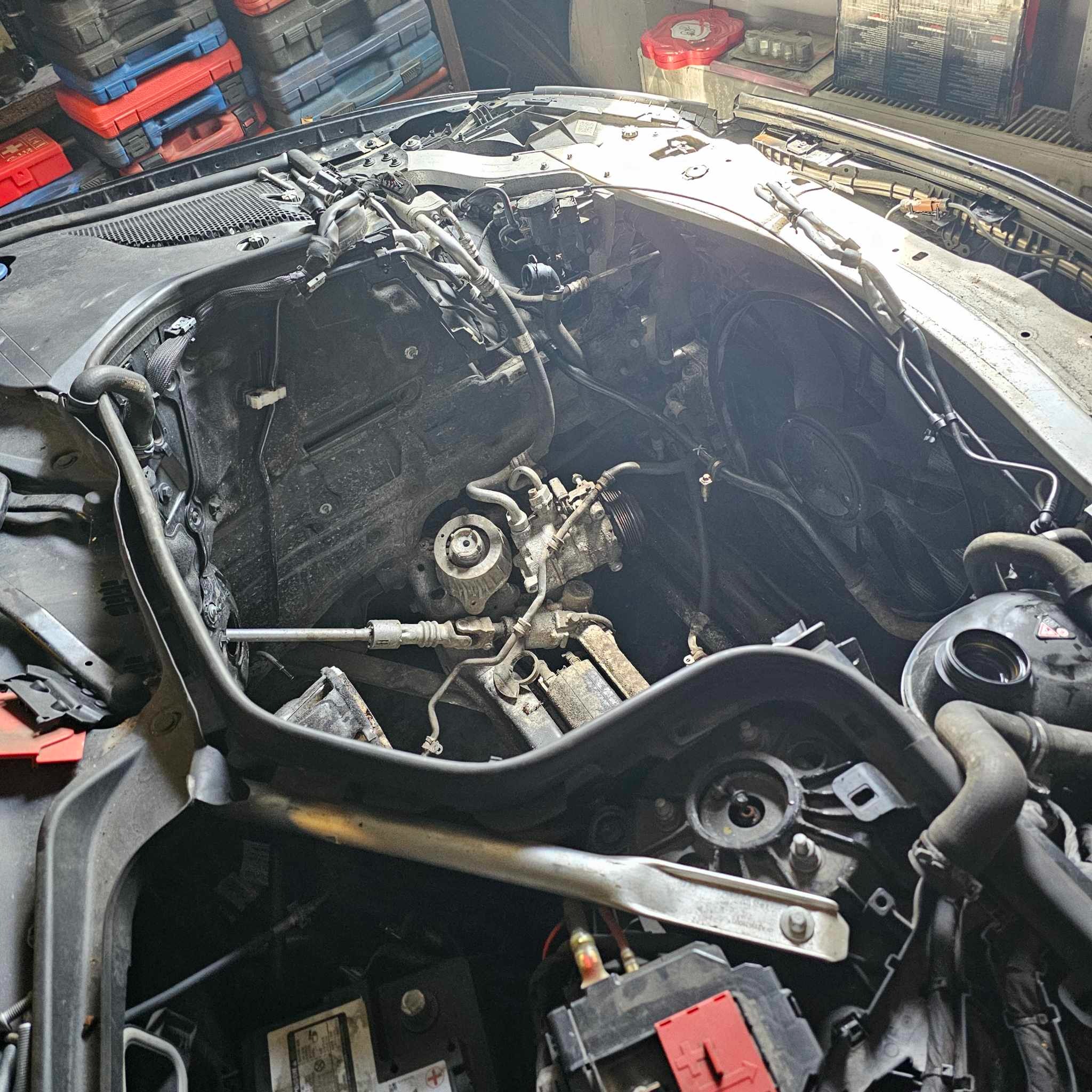 Widok z góry na otwartą komorę silnika samochodu w trakcie naprawy, widoczne elementy układu, przewody i narzędzia. Zdemontowane elementy układu chłodzenia i klimatyzacji. Akumulator widoczny...