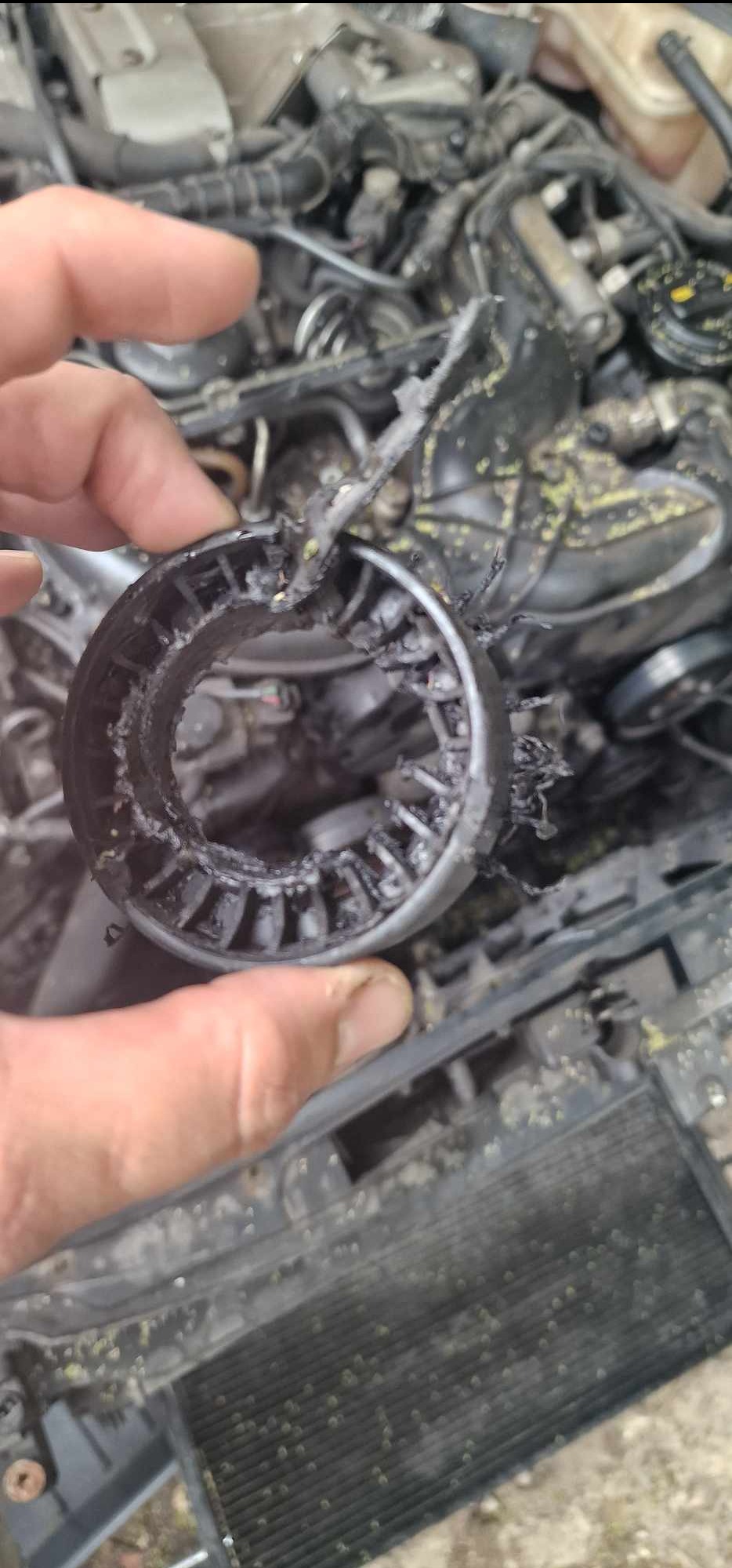 Uszkodzony, stopiony element z tworzywa sztucznego, trzymany w dłoni na tle zabrudzonego silnika samochodowego. Widoczne ślady pożaru lub wysokiej temperatury.
