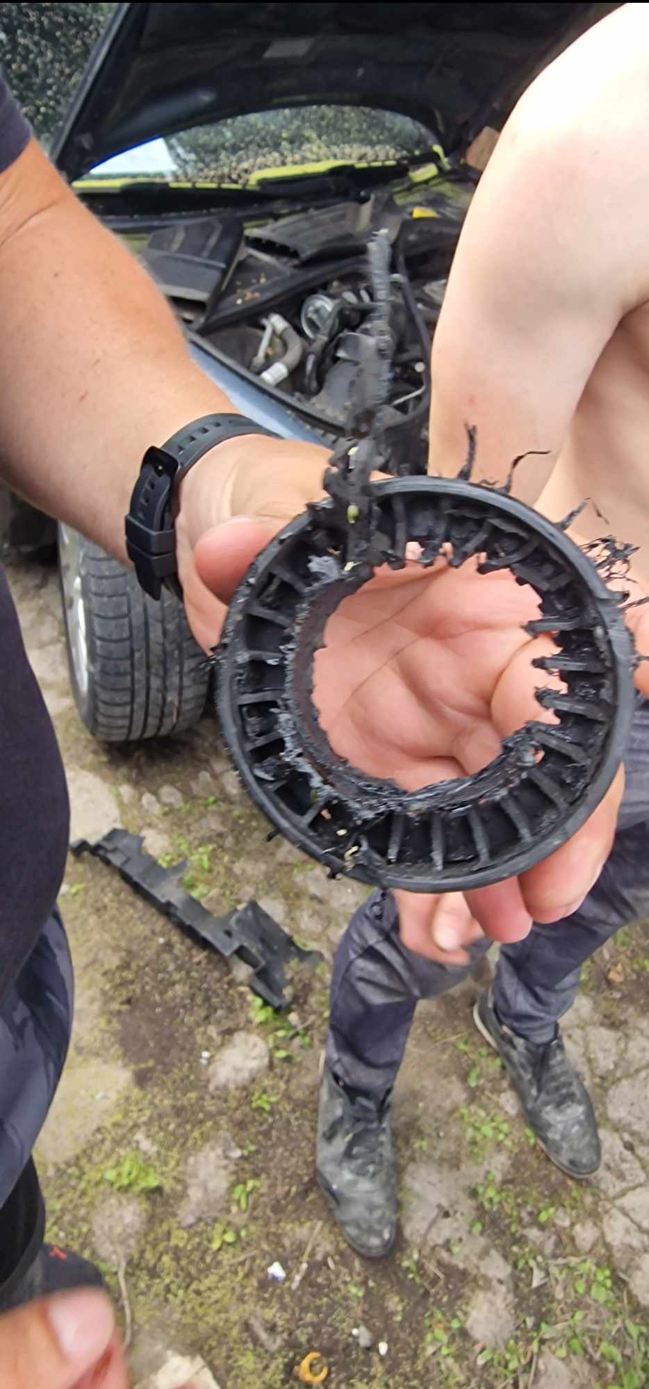 Uszkodzona, czarna, okrągła część samochodowa trzymana w dłoniach na tle otwartej maski samochodu i ziemi.