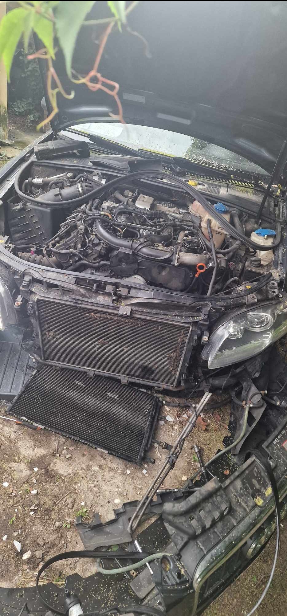 Czarny samochód z otwartą maską, widoczny silnik i zdemontowany chłodnica, elementy karoserii leżą na ziemi.