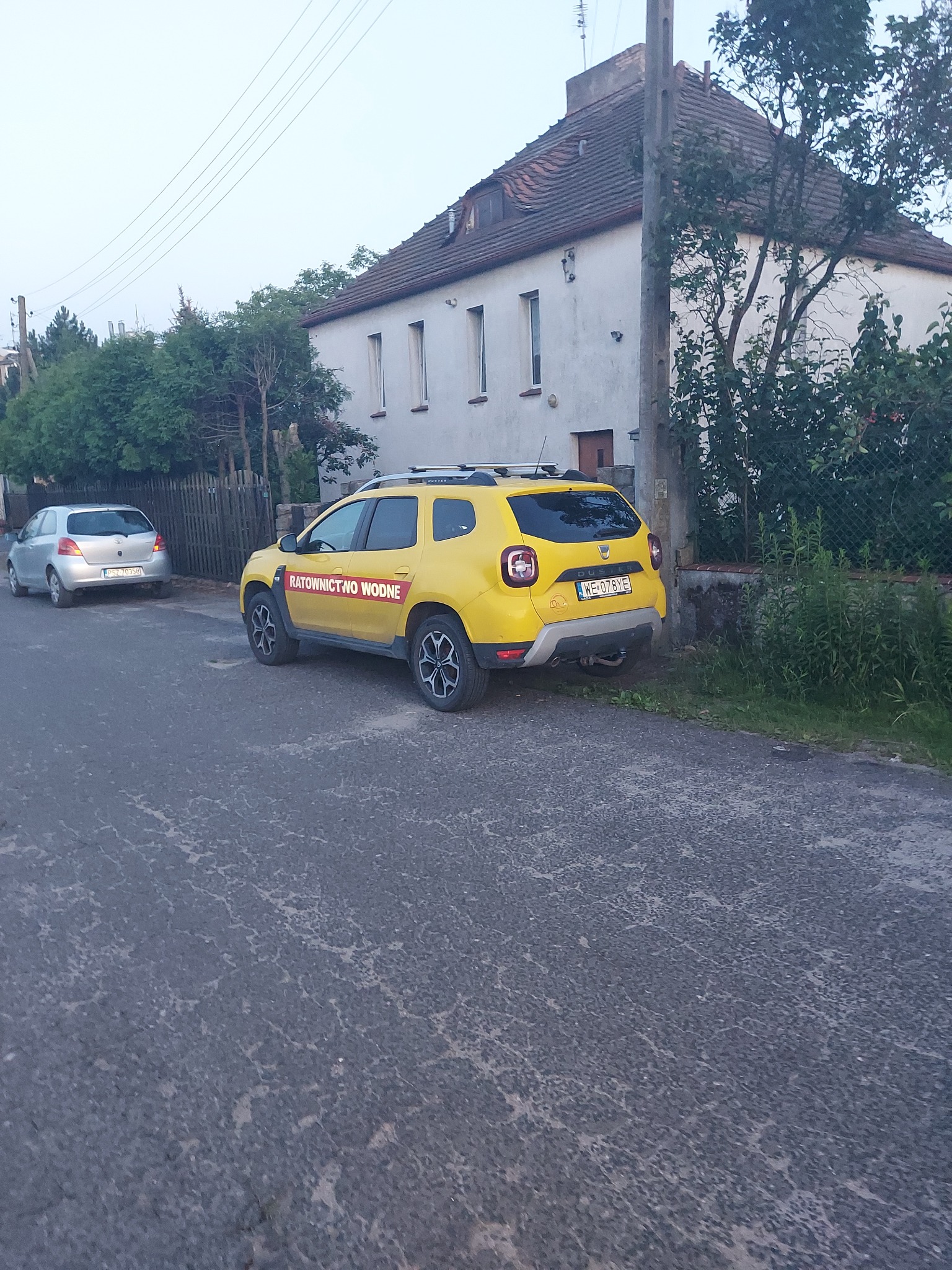 Żółty samochód z napisem 'Ratownictwo Wodne' zaparkowany na ulicy, w tle budynek mieszkalny i zieleń.