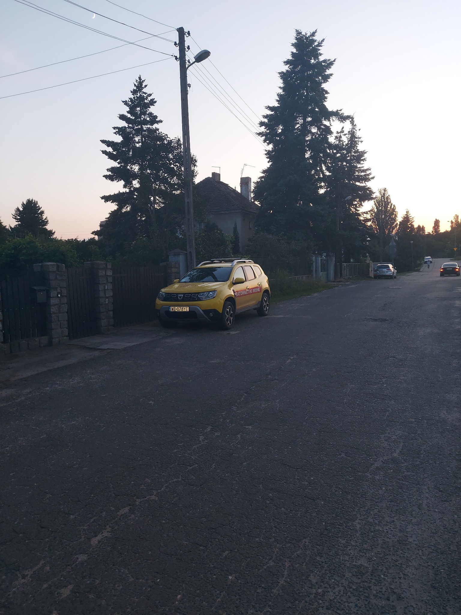 Żółty samochód osobowy zaparkowany przy ulicy w pobliżu ogrodzenia i drzew, widoczne inne pojazdy w oddali.