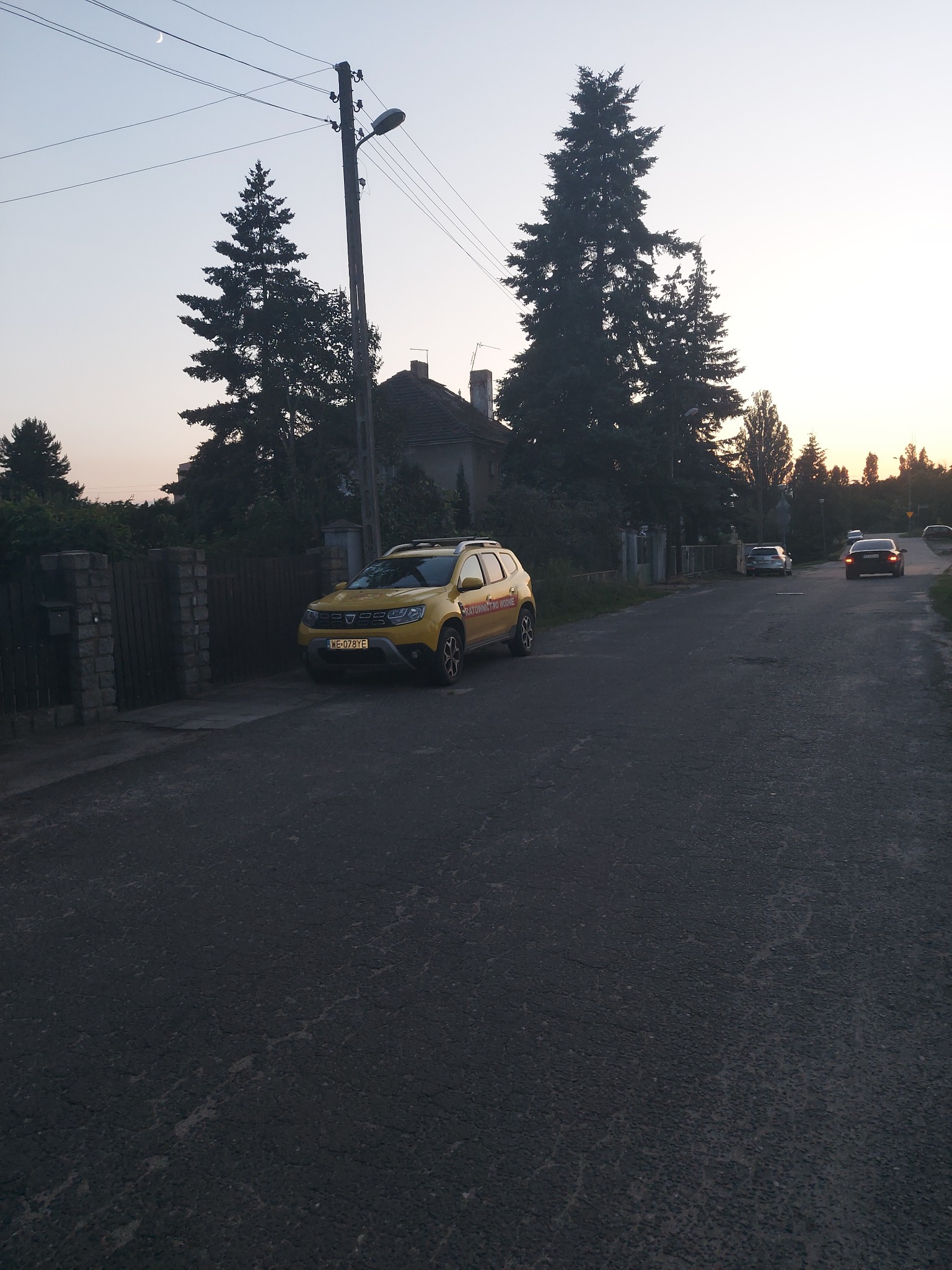 Żółty samochód Dacia Duster zaparkowany na poboczu drogi w cichej, wiejskiej okolicy, z widocznym księżycem w tle i innymi samochodami w oddali.