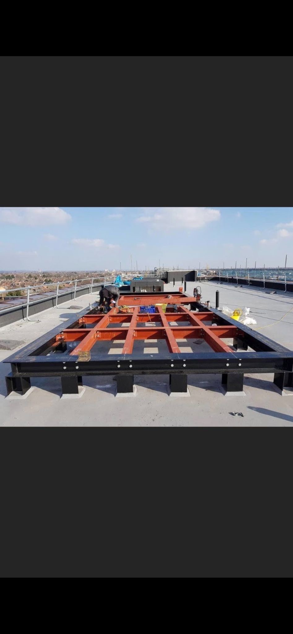 Metalowa konstrukcja szkieletowa w kolorze czarnym i pomarańczowym, prawdopodobnie pod budowę hali, montowana na dachu budynku z widokiem na miasto w tle.