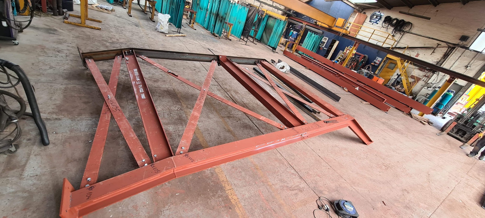 Czerwona stalowa konstrukcja kratowa z oznaczeniami leżąca na podłodze hali produkcyjnej, w tle widoczne narzędzia, butle z gazem i suwnica.