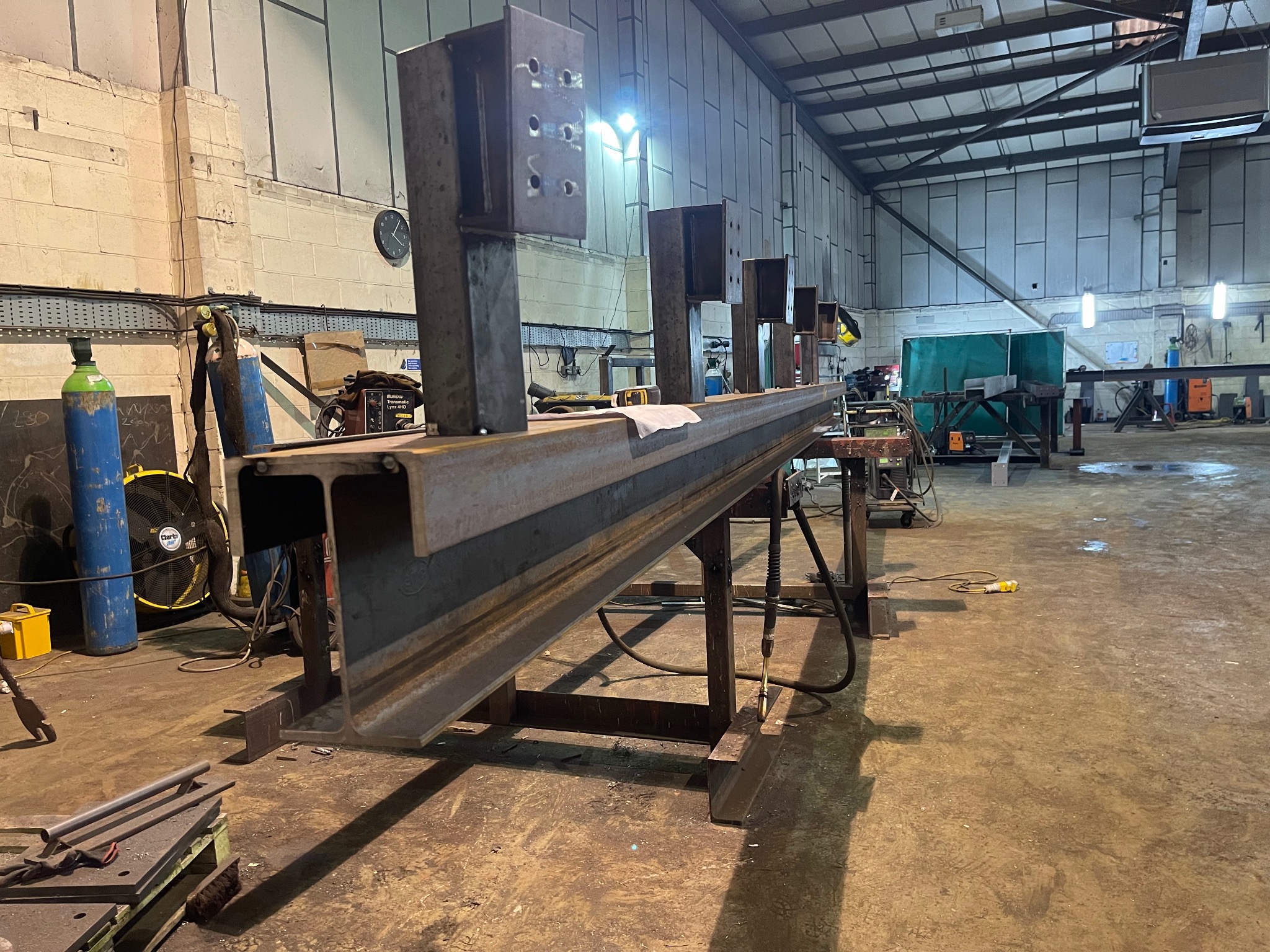 Stalowa belka dwuteowa z przyspawanymi pionowymi elementami z otworami, ustawiona na kozłach w hali produkcyjnej, widoczne butle z gazem i sprzęt spawalniczy w tle.