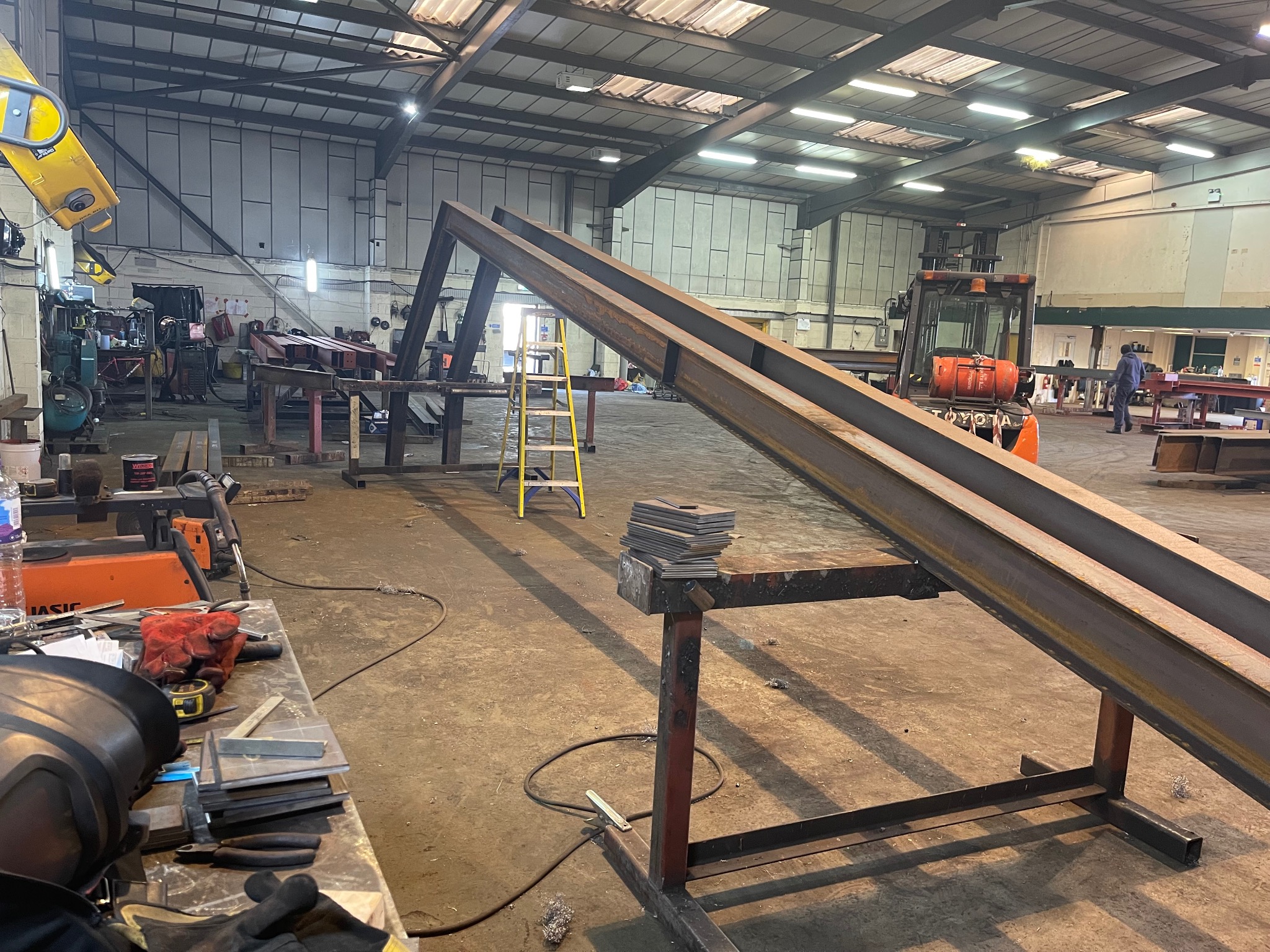 Wnętrze hali produkcyjnej z elementami konstrukcji stalowej w trakcie montażu, widoczne profile stalowe na kozłach, spawarka, narzędzia i drabina.