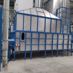 Apanotech - Złożona przemysłowa konstrukcja metalowa z niebieską ramą i srebrnymi panelami, prawdopodobnie urządzenie do obróbki powierzchni lub kabina lakiernicza, z widocznymi przewodami elektrycznymi...