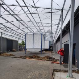 Apanotech - Montaż konstrukcji stalowej hali z silosami, widoczny pracownik w czerwonej bluzie i szarych spodniach, teren budowy z rozkopaną kostką brukową i stertami ziemi.