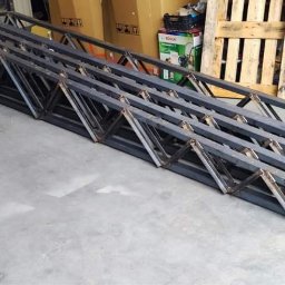 Apanotech - Długie, spawane konstrukcje kratownicowe z metalowych profili, ułożone równolegle na betonowej podłodze w hali produkcyjnej, widoczne ślady spawania i obróbki.