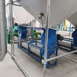 Instalacja przemysłowa z wentylatorami i rurami wentylacyjnymi, widoczne elementy metalowe i białe worki filtracyjne, w tle wózek widłowy.