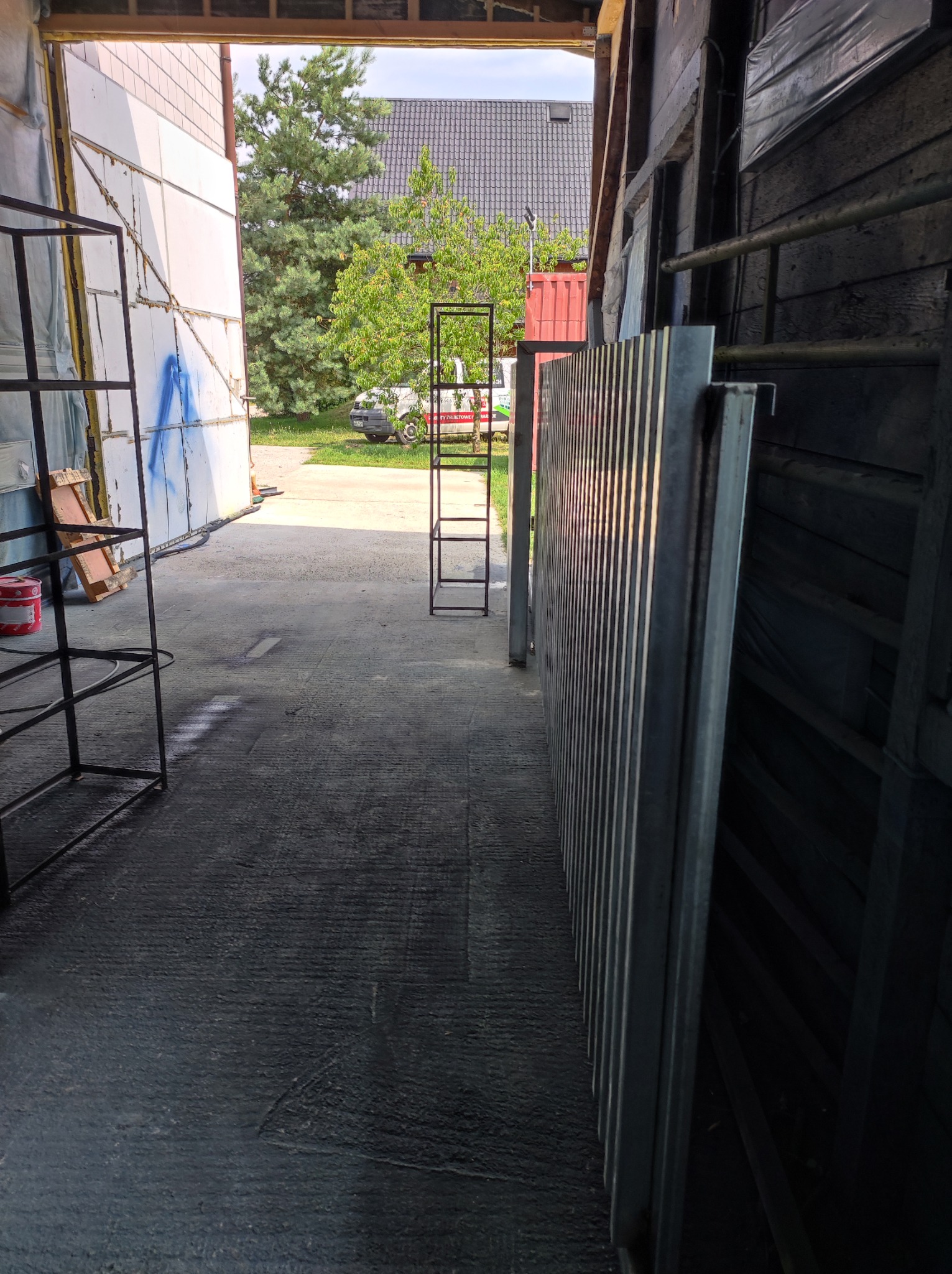 Metalowe konstrukcje ramowe, prawdopodobnie regały, w warsztacie lub hali produkcyjnej. Część konstrukcji stoi na zewnątrz, widoczny budynek i zieleń w tle.