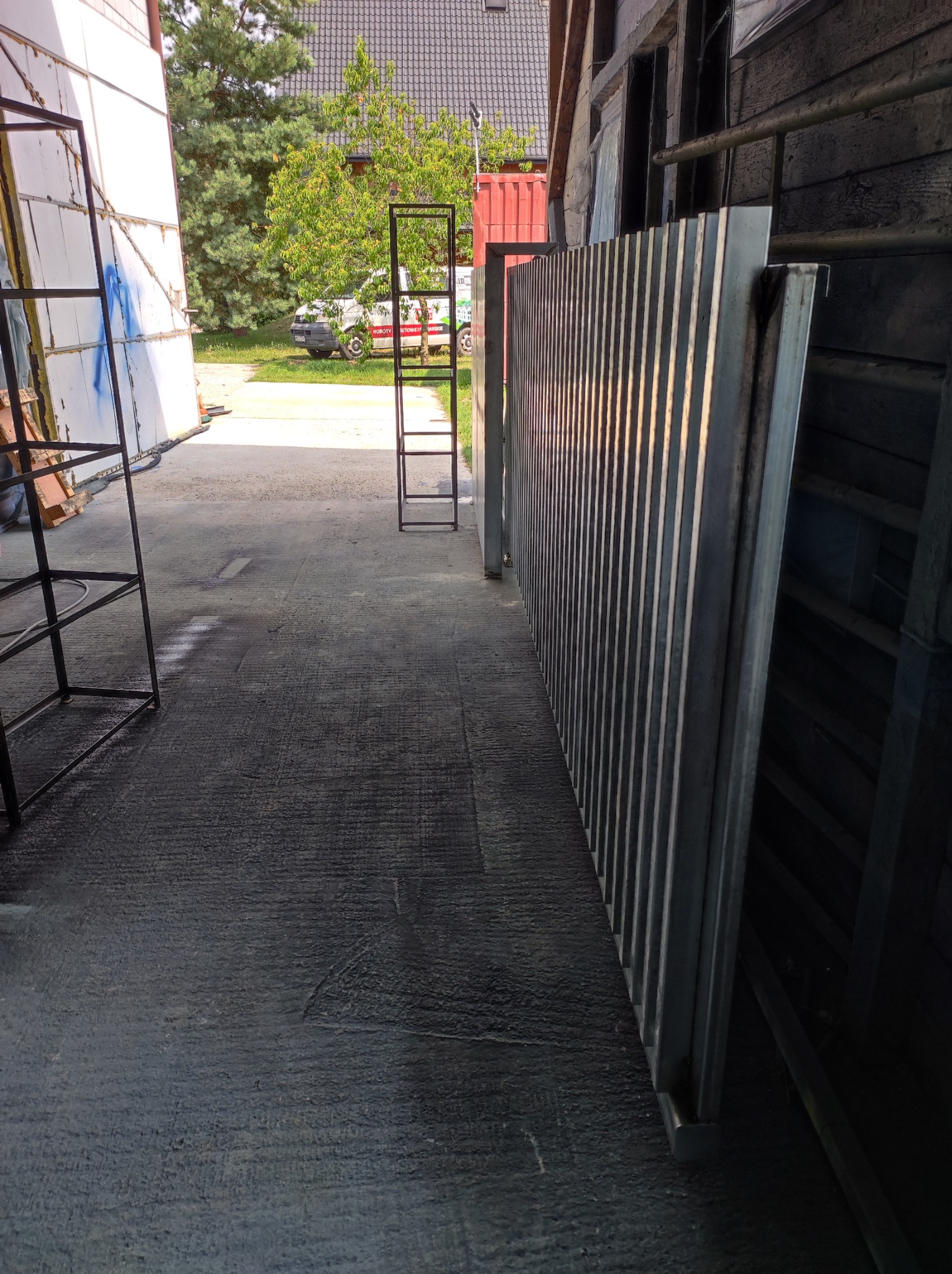 Stalowe elementy konstrukcyjne, prawdopodobnie kraty lub panele, ustawione pionowo w warsztacie lub hali produkcyjnej, z widocznym fragmentem podwórza i samochodem firmy w tle.