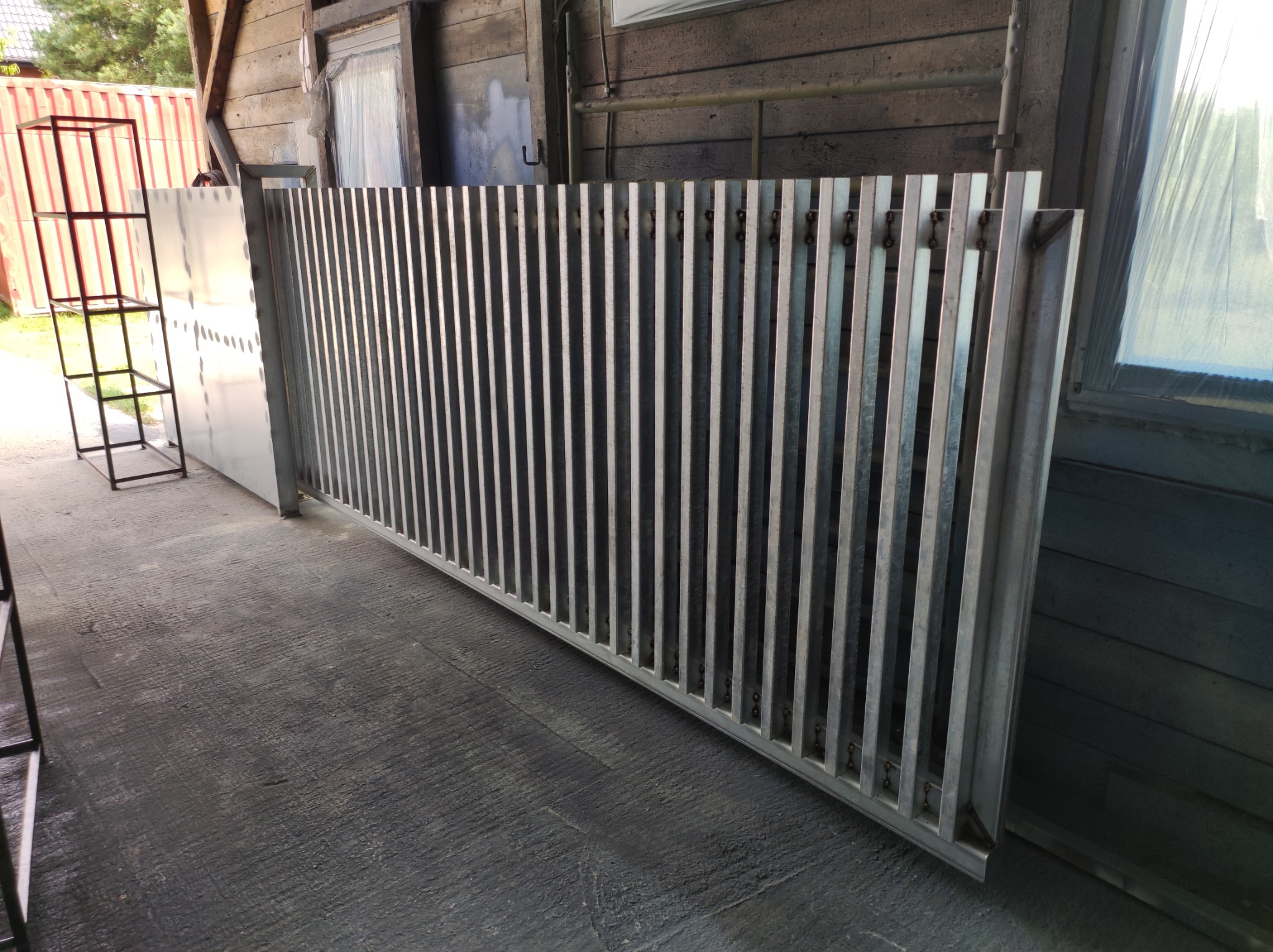 Metalowa brama z pionowymi szczeblami, surowe wykończenie, widoczne spawy, ustawiona w warsztacie na betonowej posadzce, ściana z desek w tle.