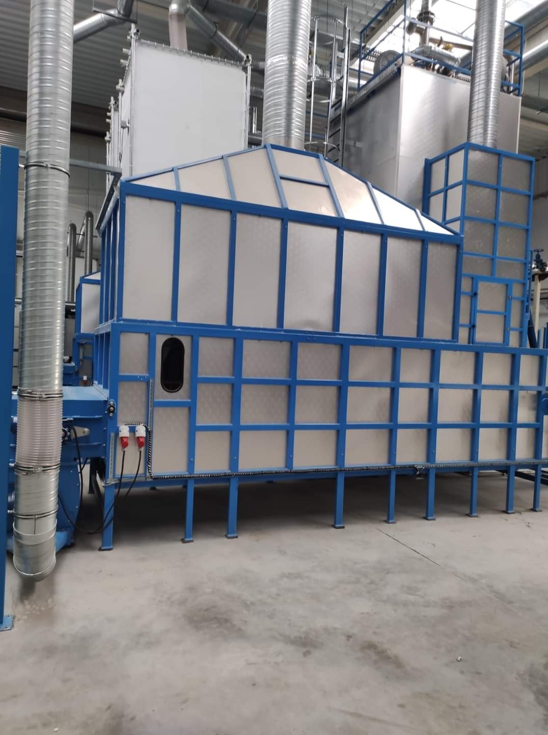 Złożona przemysłowa konstrukcja metalowa z niebieską ramą i srebrnymi panelami, prawdopodobnie urządzenie do obróbki powierzchni lub kabina lakiernicza, z widocznymi przewodami elektrycznymi...