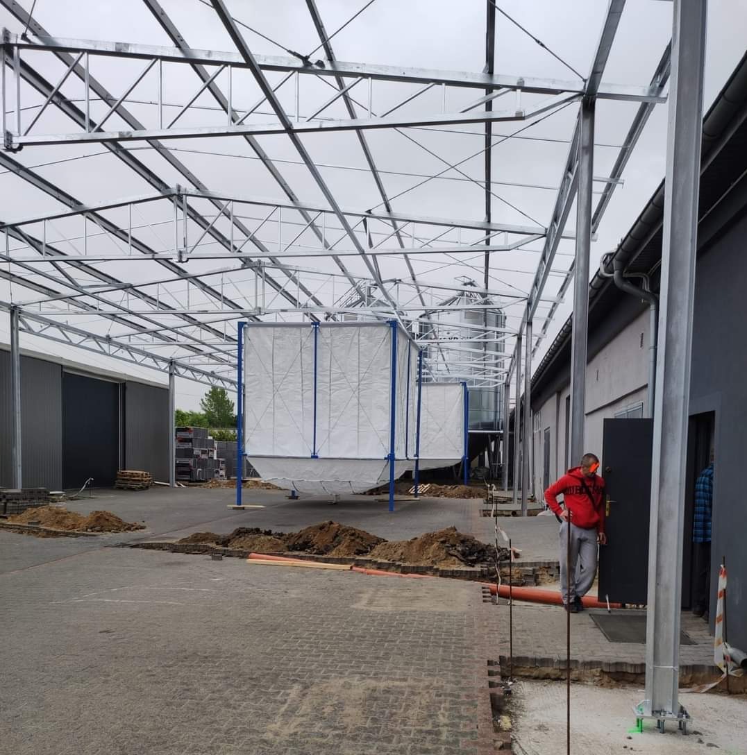 Montaż konstrukcji stalowej hali z silosami, widoczny pracownik w czerwonej bluzie i szarych spodniach, teren budowy z rozkopaną kostką brukową i stertami ziemi.