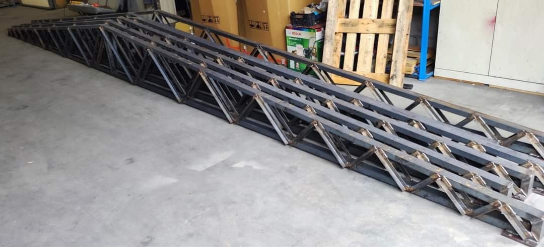 Długie, spawane konstrukcje kratownicowe z metalowych profili, ułożone równolegle na betonowej podłodze w hali produkcyjnej, widoczne ślady spawania i obróbki.