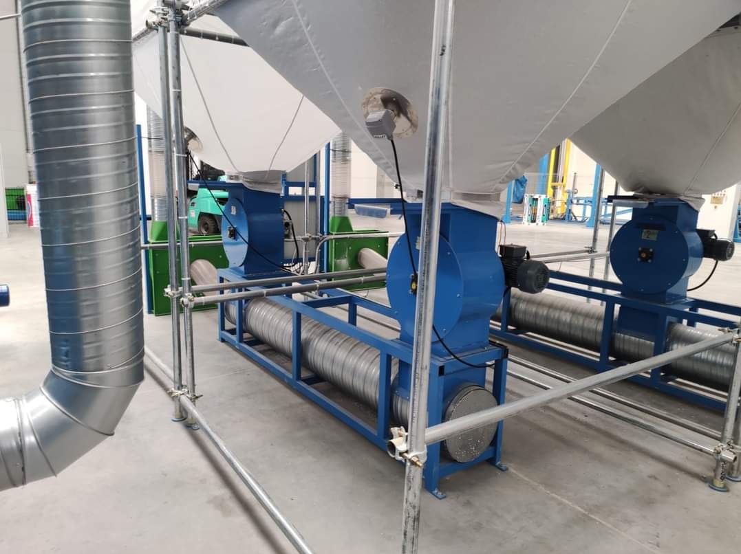 Instalacja przemysłowa z wentylatorami i rurami wentylacyjnymi, widoczne elementy metalowe i białe worki filtracyjne, w tle wózek widłowy.