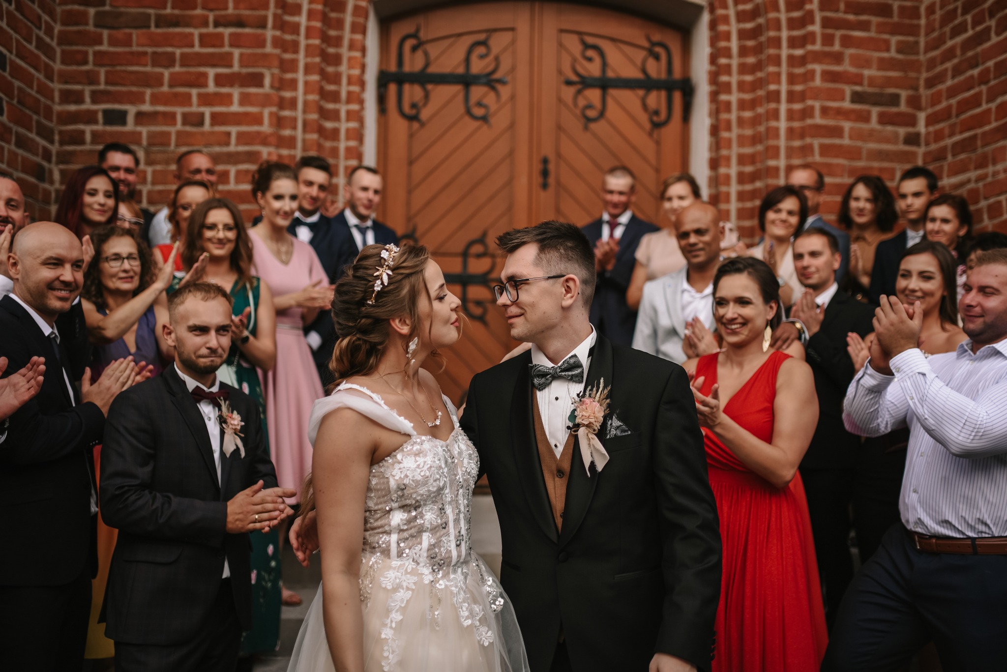 Para młoda w eleganckich strojach, otoczona gośćmi klaszczącymi przed ceglanym budynkiem z drewnianymi drzwiami.