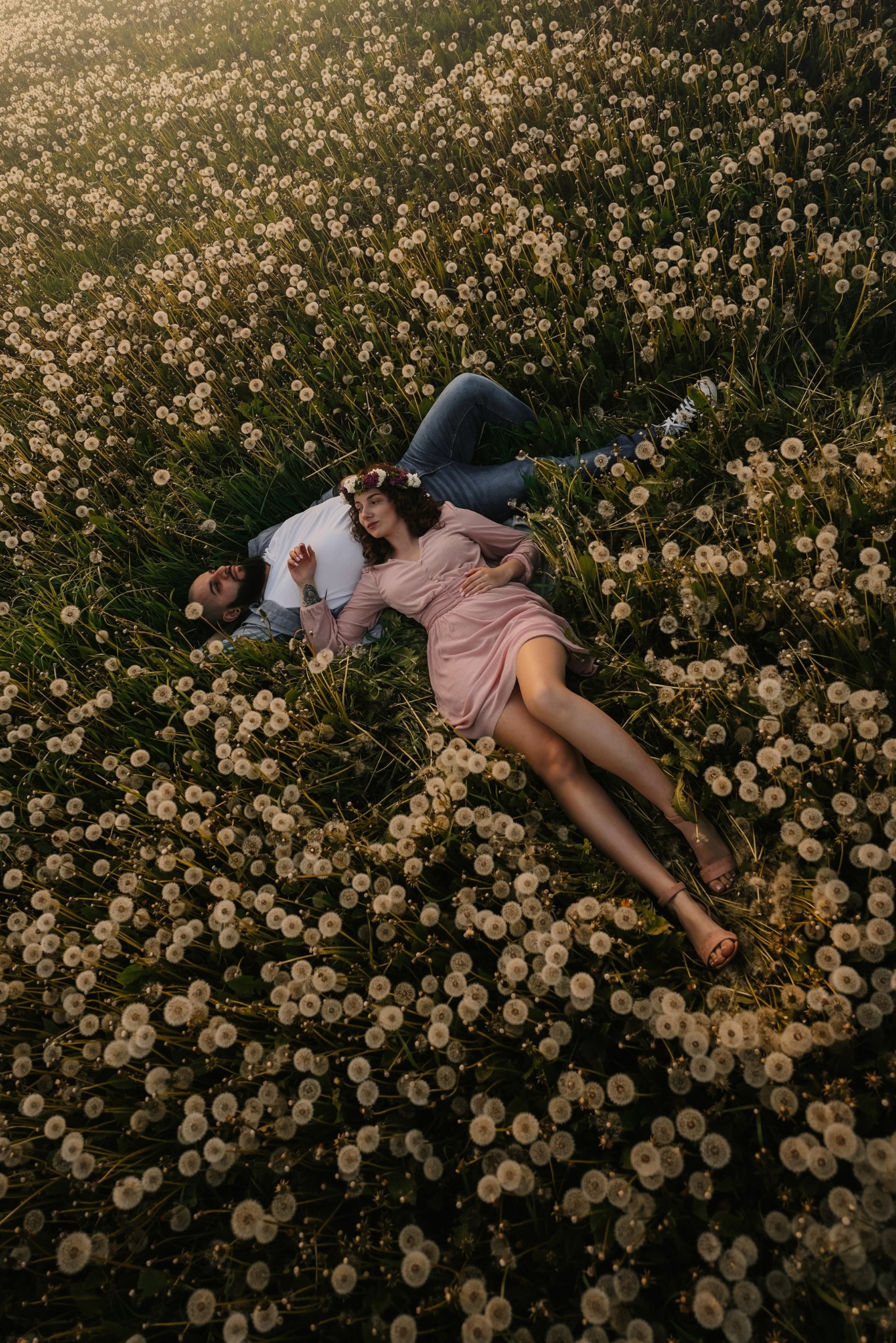 Para leży na łące dmuchawców, kobieta w różowej sukience i wianku, mężczyzna w jeansach i białej koszulce, widok z góry, ciepłe światło zachodzącego słońca.