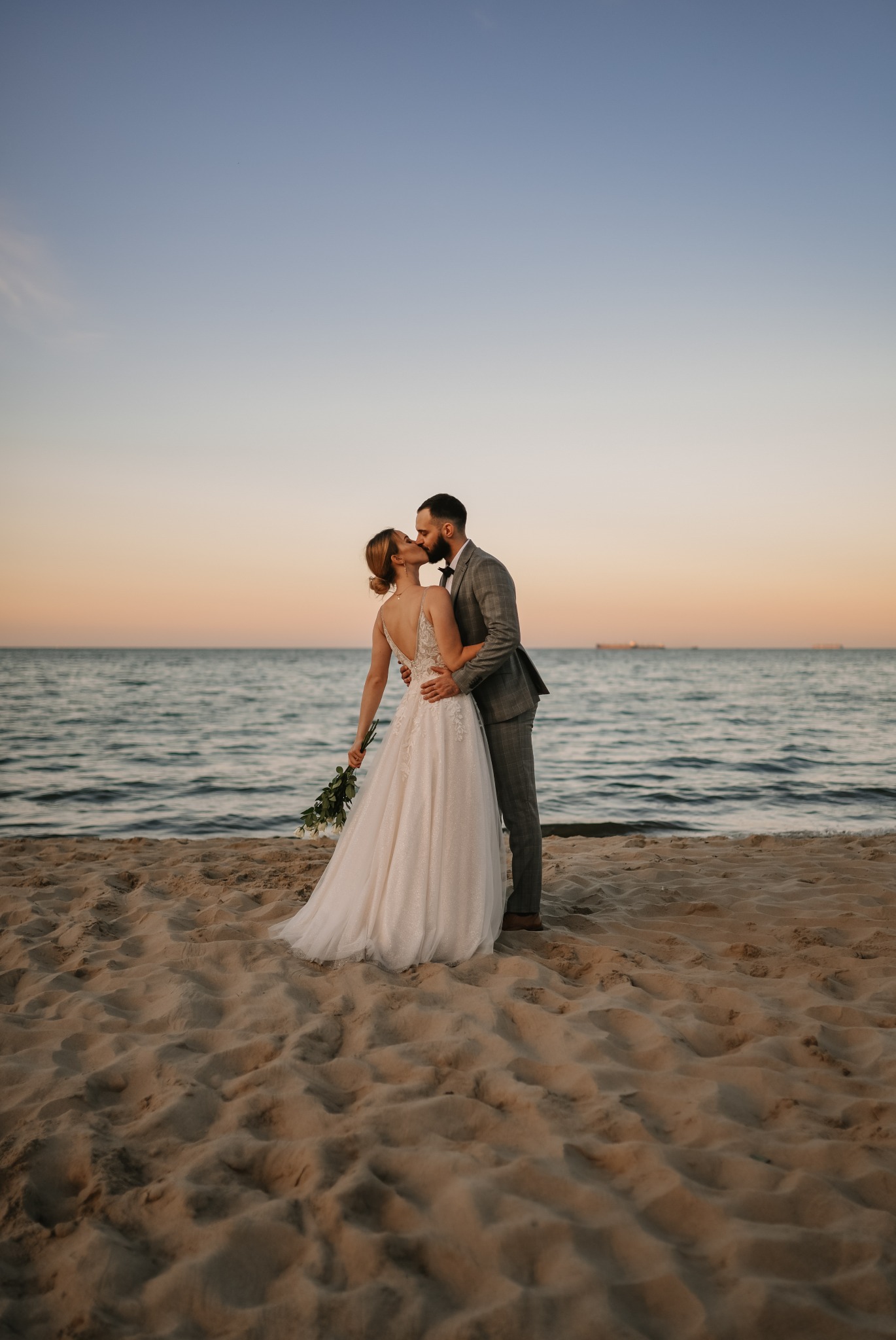 Para młoda całuje się na piaszczystej plaży o zachodzie słońca, panna młoda w sukni z odkrytymi plecami trzyma bukiet białych róż, pan młody w szarej kraciastej marynarce i muszce.