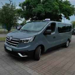 Szary van Renault Trafic z oznaczeniem Taxi na dachu, zaparkowany na brukowanym parkingu przed budynkiem.