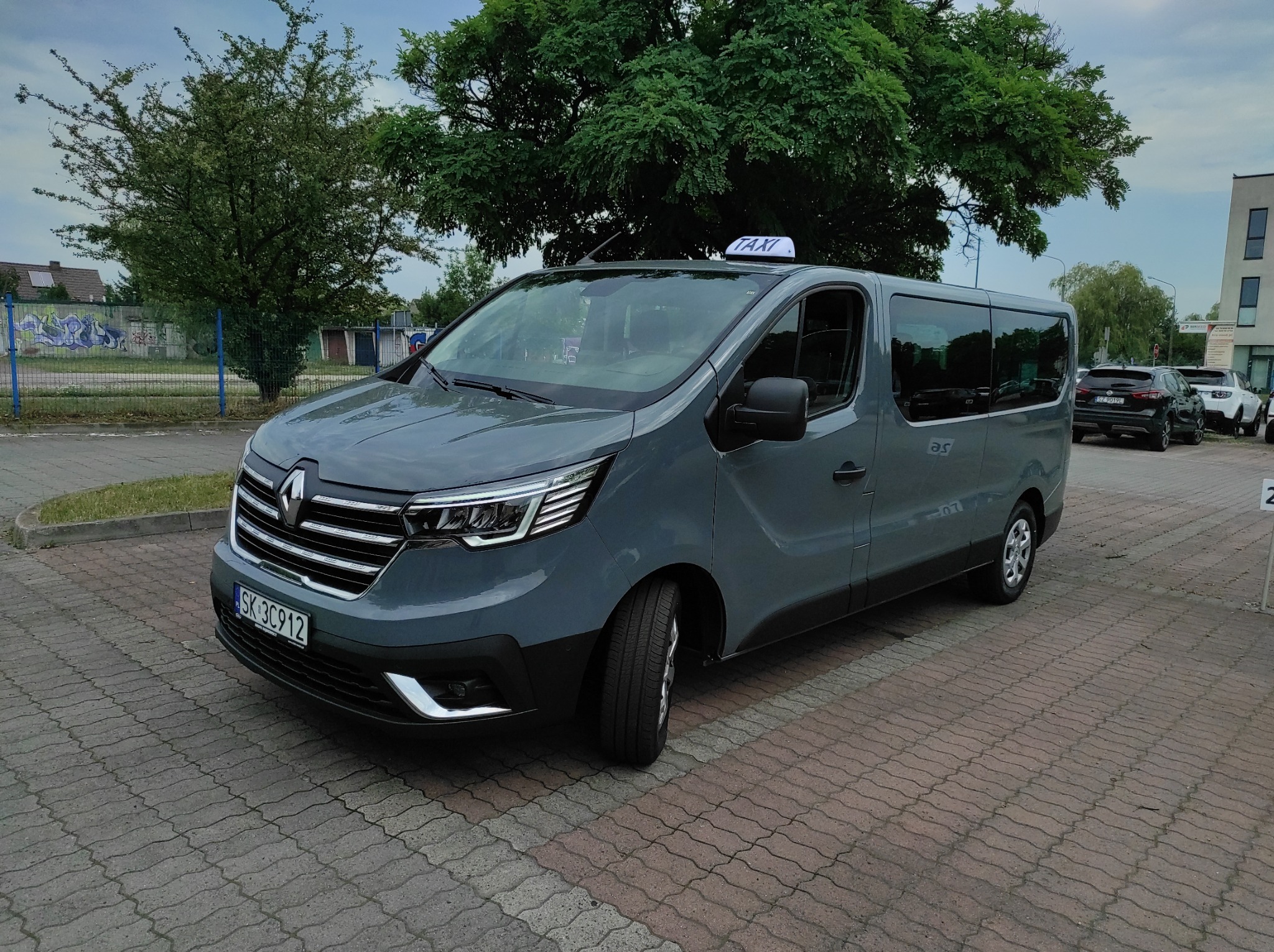 Szary van Renault Trafic z oznaczeniem Taxi na dachu, zaparkowany na brukowanym parkingu przed budynkiem.